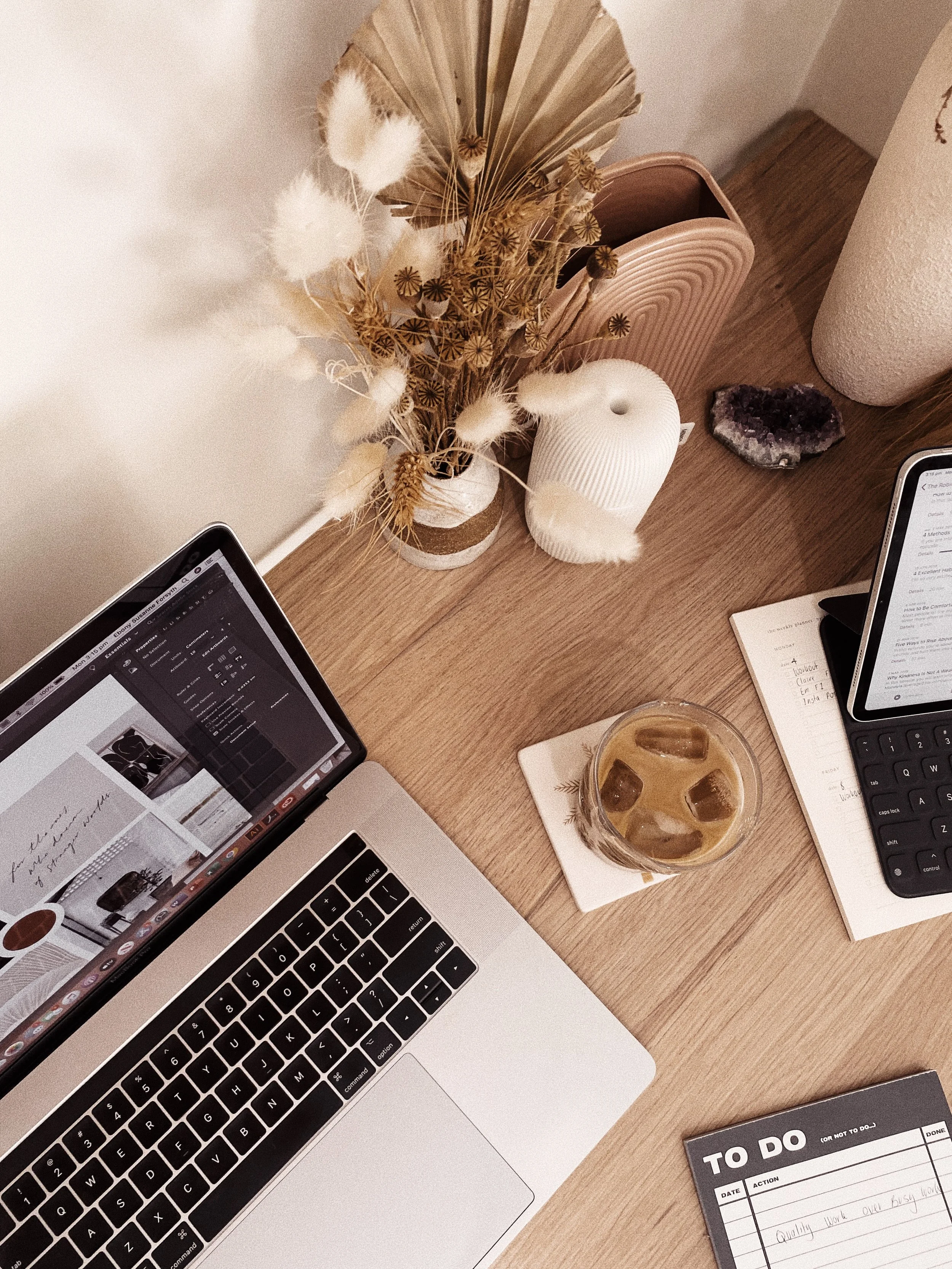 A workspace desk with a MacBook laptop, a cellphone, a glass of iced coffee, a notebook, a keyboard, and decorative items including a dried flower arrangement, a geode, and vases.