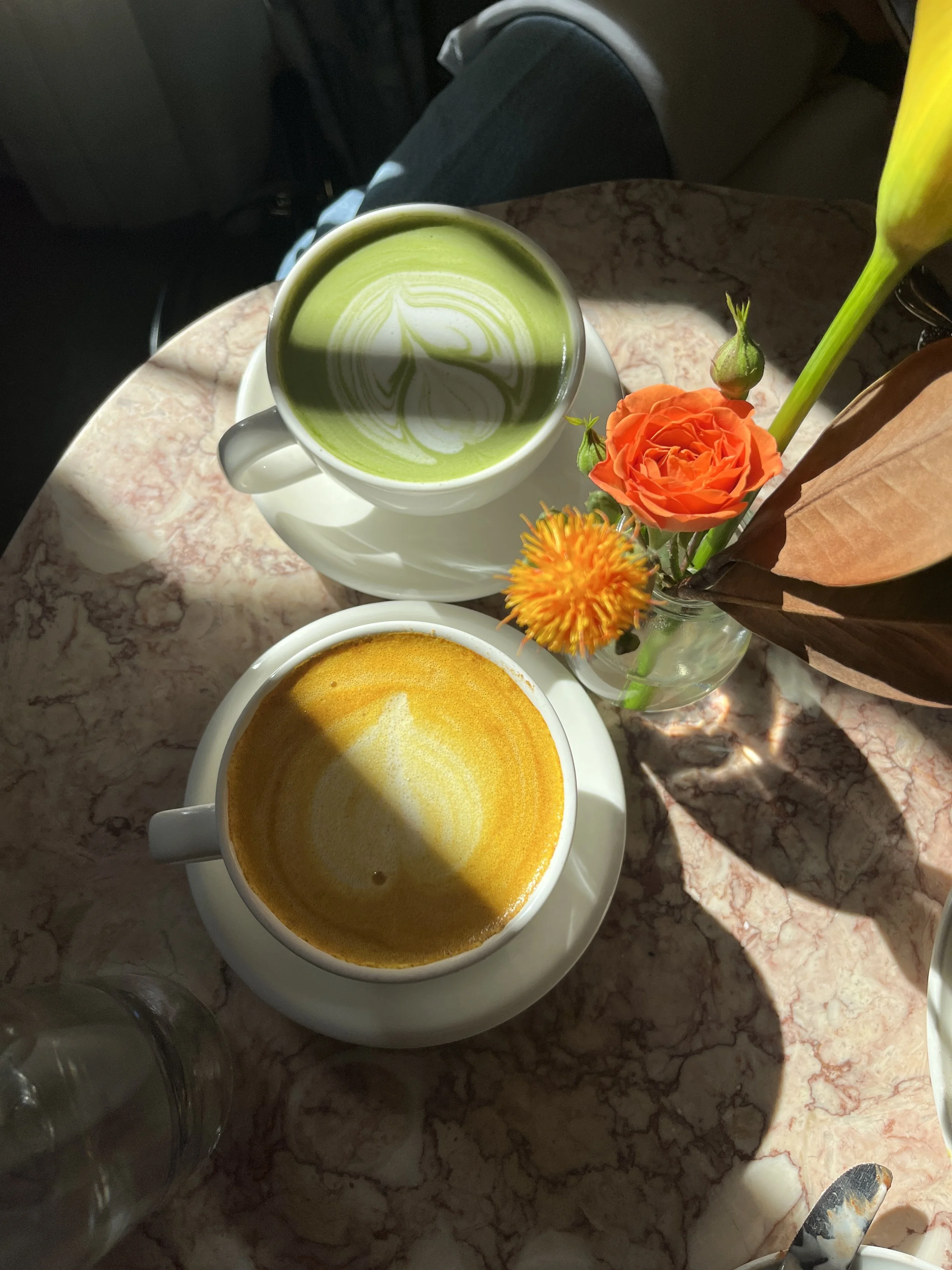Top-down view of a table with two cups of coffee, one with a green matcha latte with latte art and the other with a yellow turmeric latte, a small vase with orange and yellow flowers, and a glass of water, set on a pink and white marble surface.
