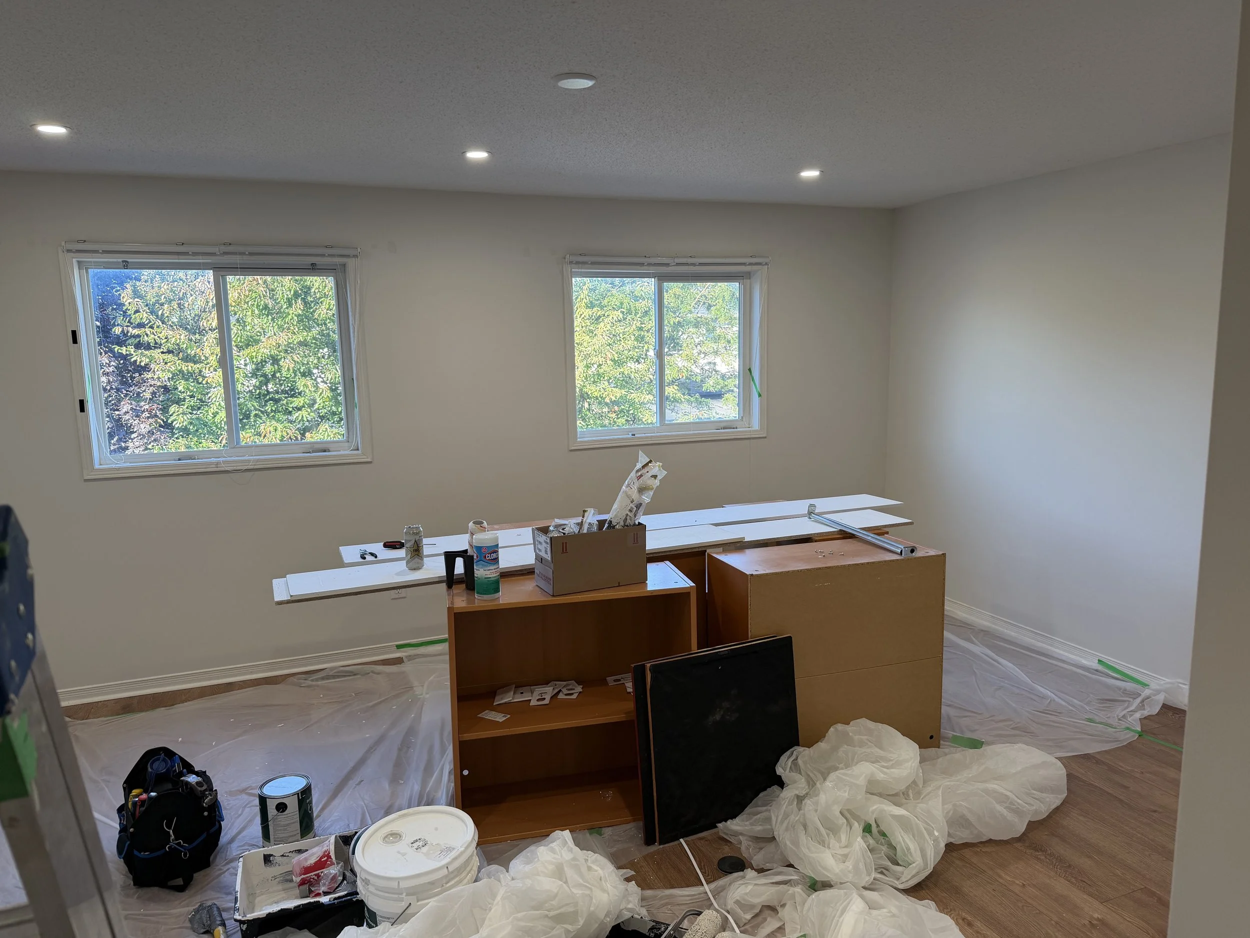Room under renovation with two windows and white walls. Construction supplies, a cabinet, and protective plastic sheeting on the floor.