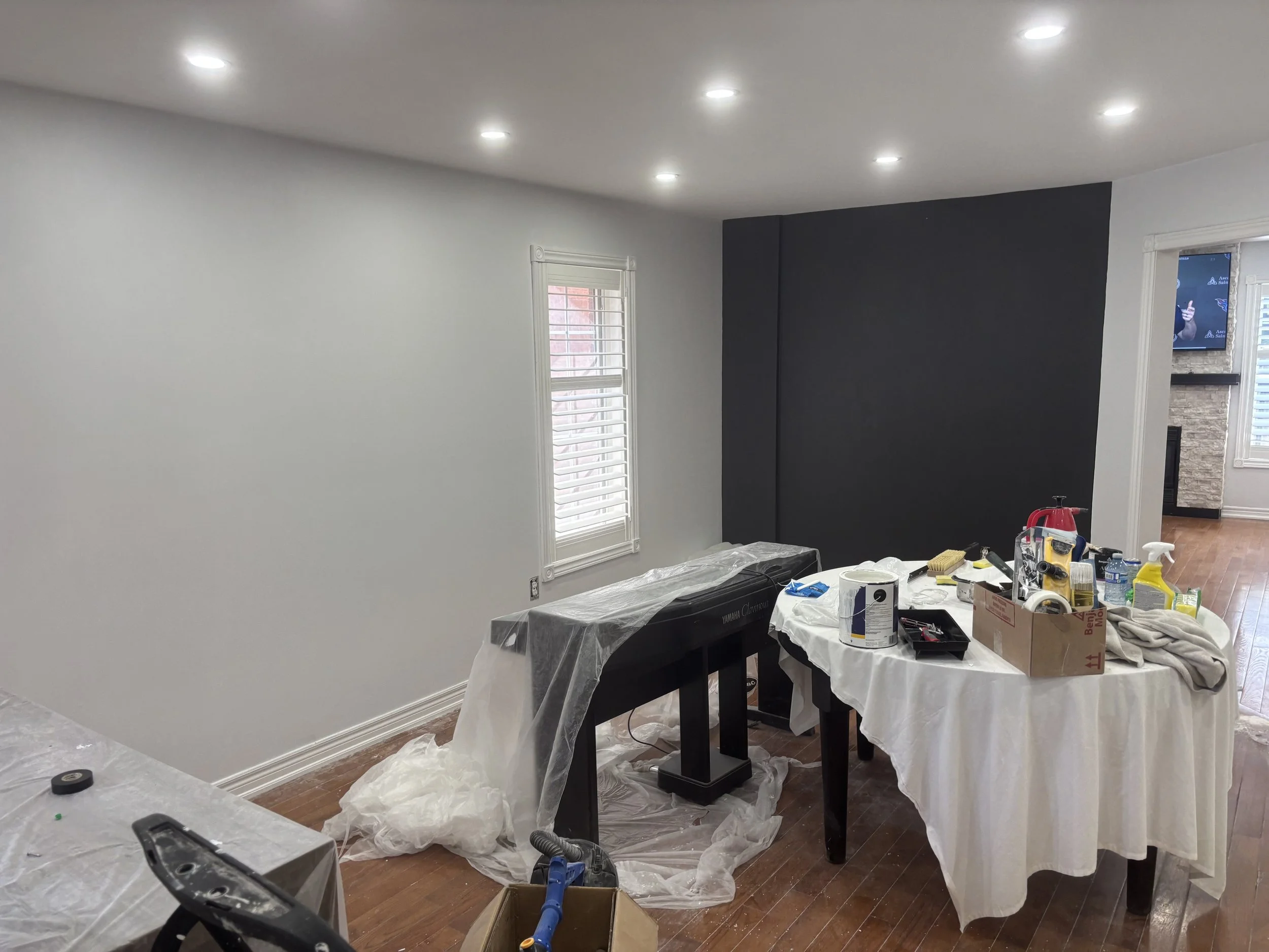 Room undergoing renovation with white and dark gray walls, ceiling lights, window with shutters, and a table covered with cleaning and painting supplies such as paint cans, brushes, spray bottles, and trash.