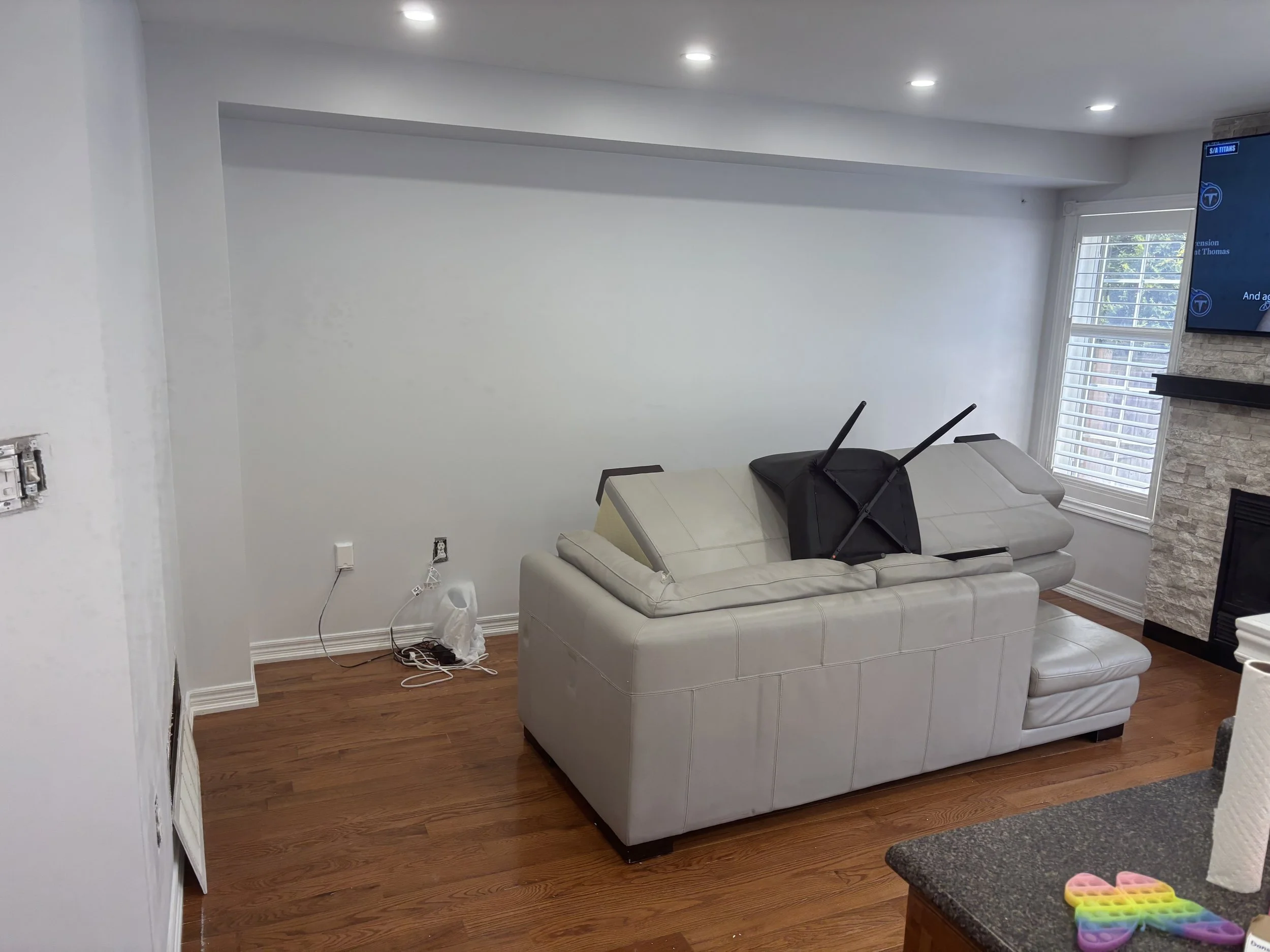 Living room with a white sofa, a wall-mounted TV, a window with white blinds, and a stone fireplace. Part of a kitchen counter with a colorful silicone ice tray is visible in the foreground.