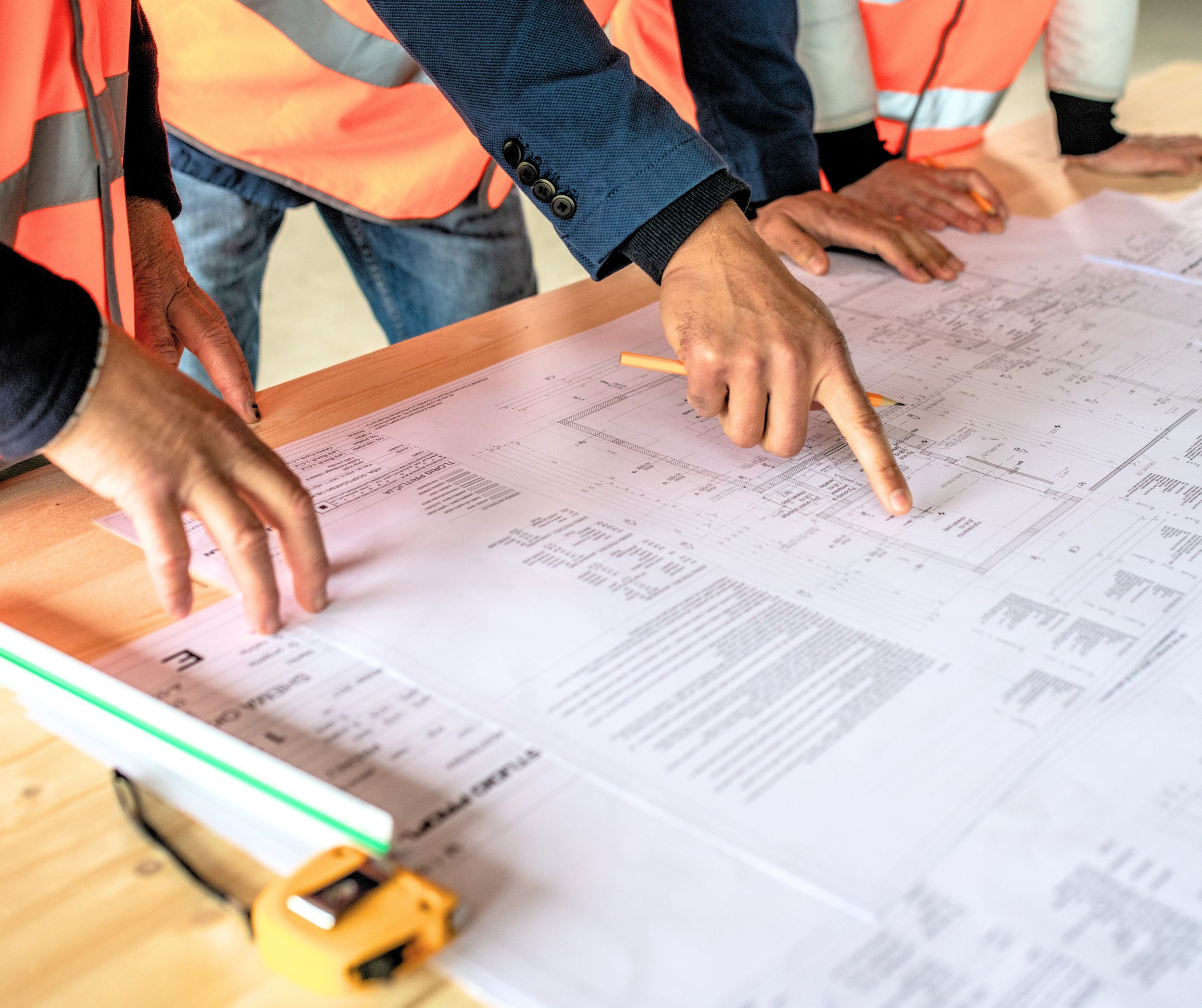 Des mains pointant des plans d'architecture ou des dessins techniques posés sur une table en bois, portés par des personnes en gilets de sécurité oranges.
