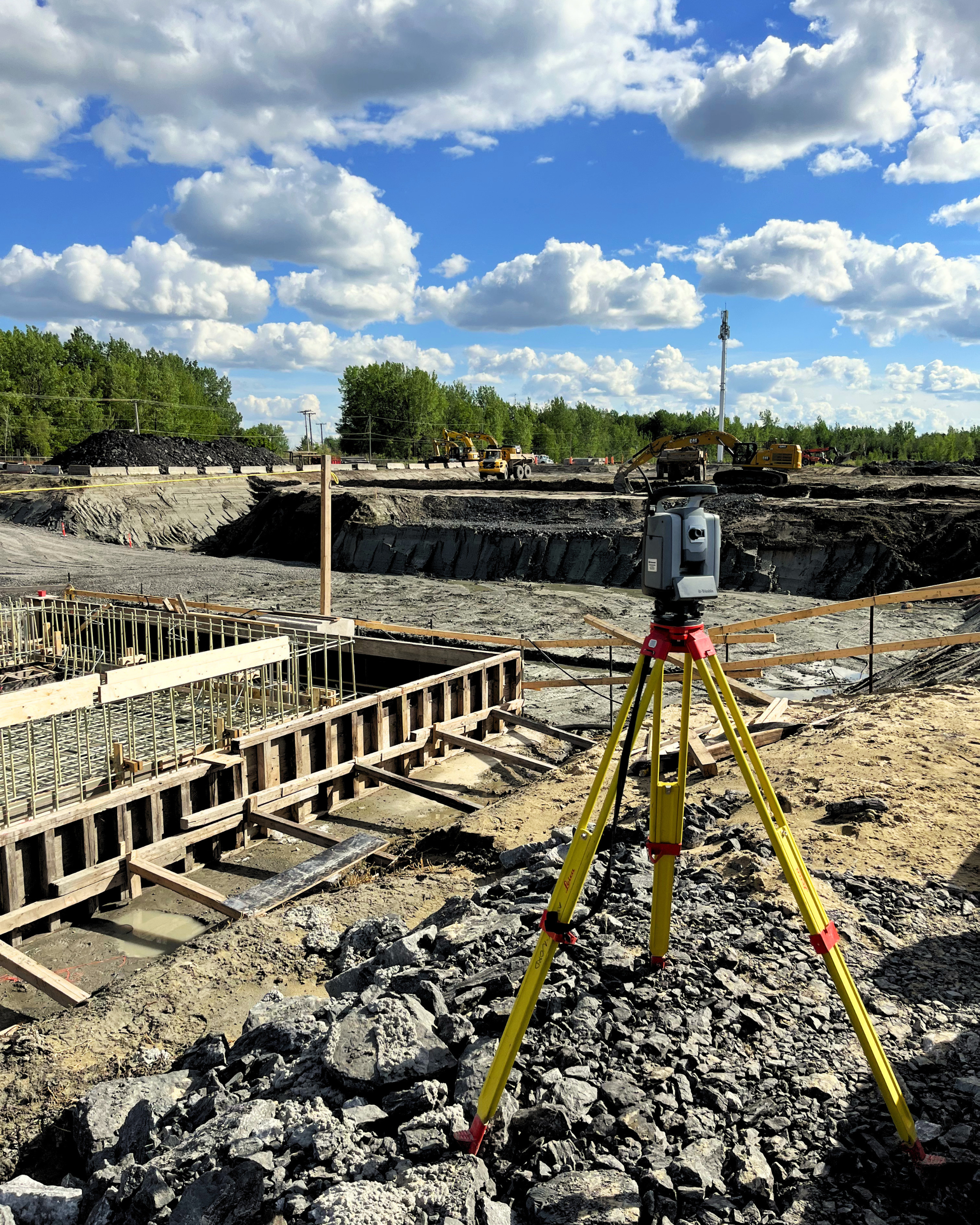 Une zone de construction en plein air avec des excavatrices jaunes en activité, un niveau laser jaune sur un trépied en avant-plan, des roches et des excavations profondes, un ciel bleu avec des nuages blancs, et des arbres en arrière-plan.