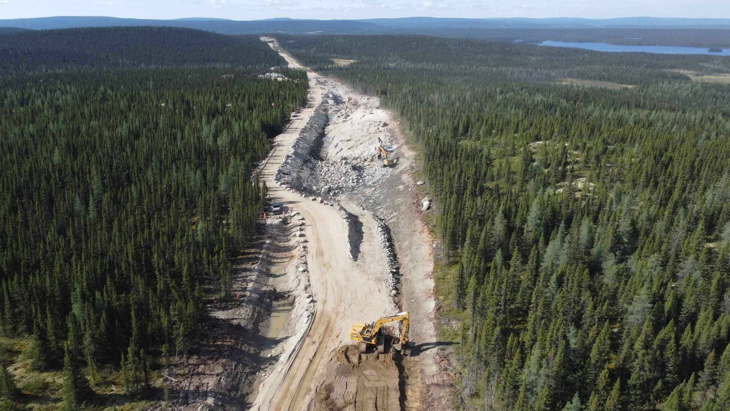 Travaux de construction de route dans une forêt dense, avec des excavatrices et des véhicules, au milieu d'une zone boisée avec un lac visible au loin.