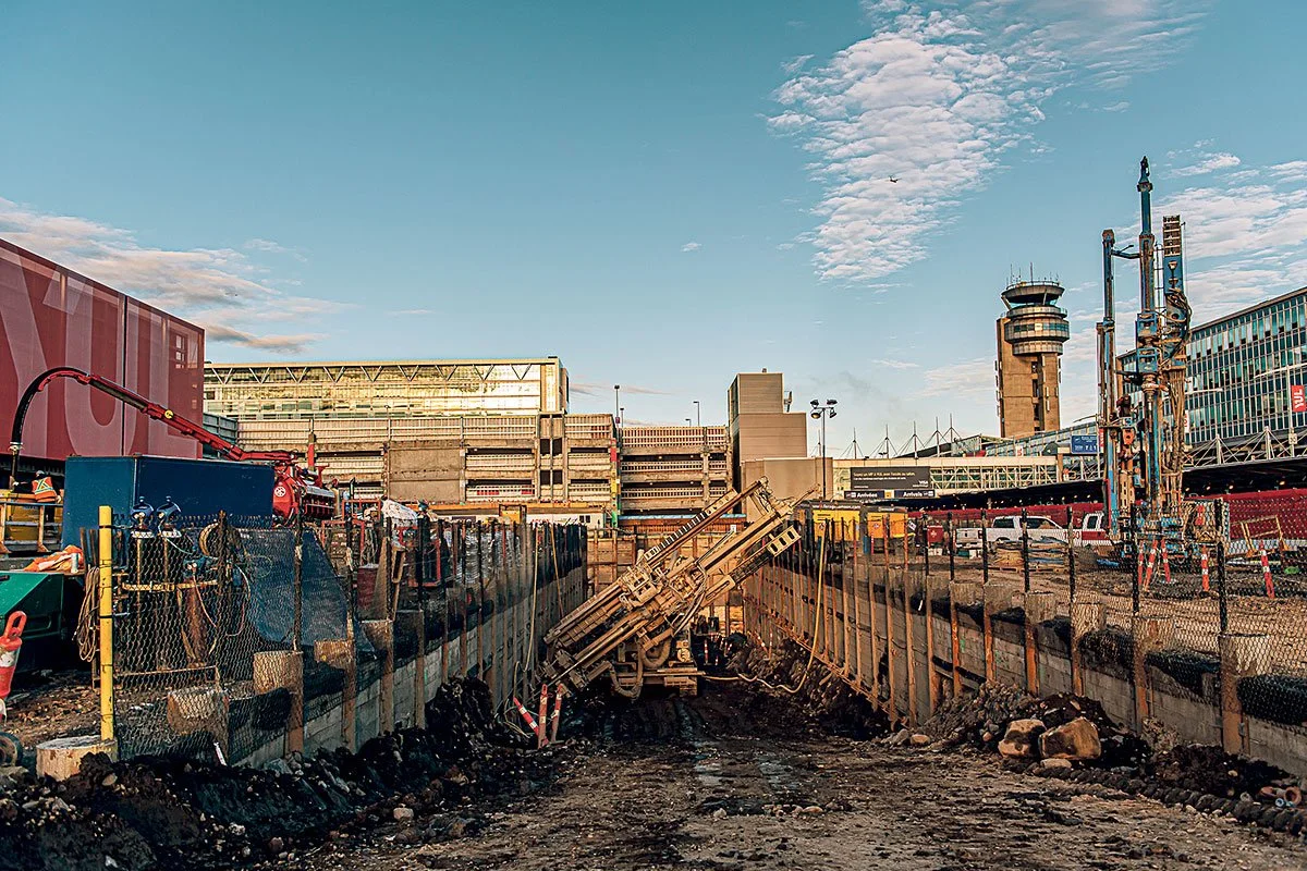 Travail de construction ou de rénovation urbaine dans une zone de chantier avec des excavations et des machines, le tout dans un paysage urbain avec des bâtiments modernes et un ciel partiellement nuageux.