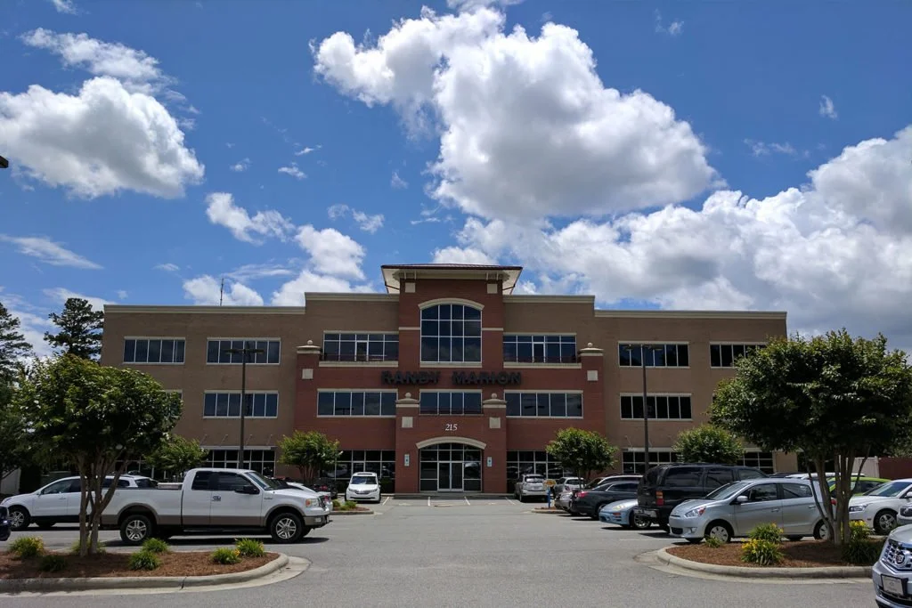 A multi-story office building with a parking lot in front, trees along the perimeter, and a partly cloudy sky overhead.