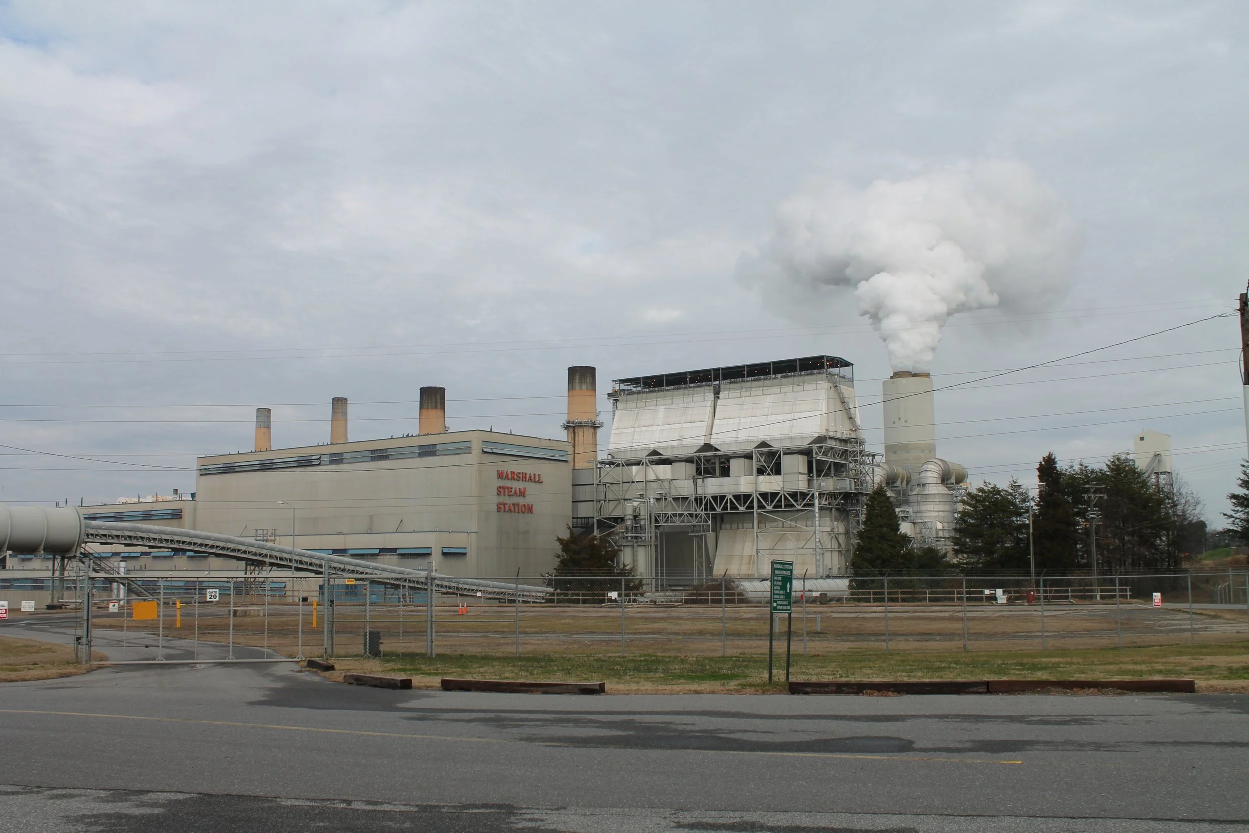 A large industrial power plant with multiple smokestacks emitting white smoke, with a sign that reads 'Marshall Steam Station' and a chain-link fence in the foreground.