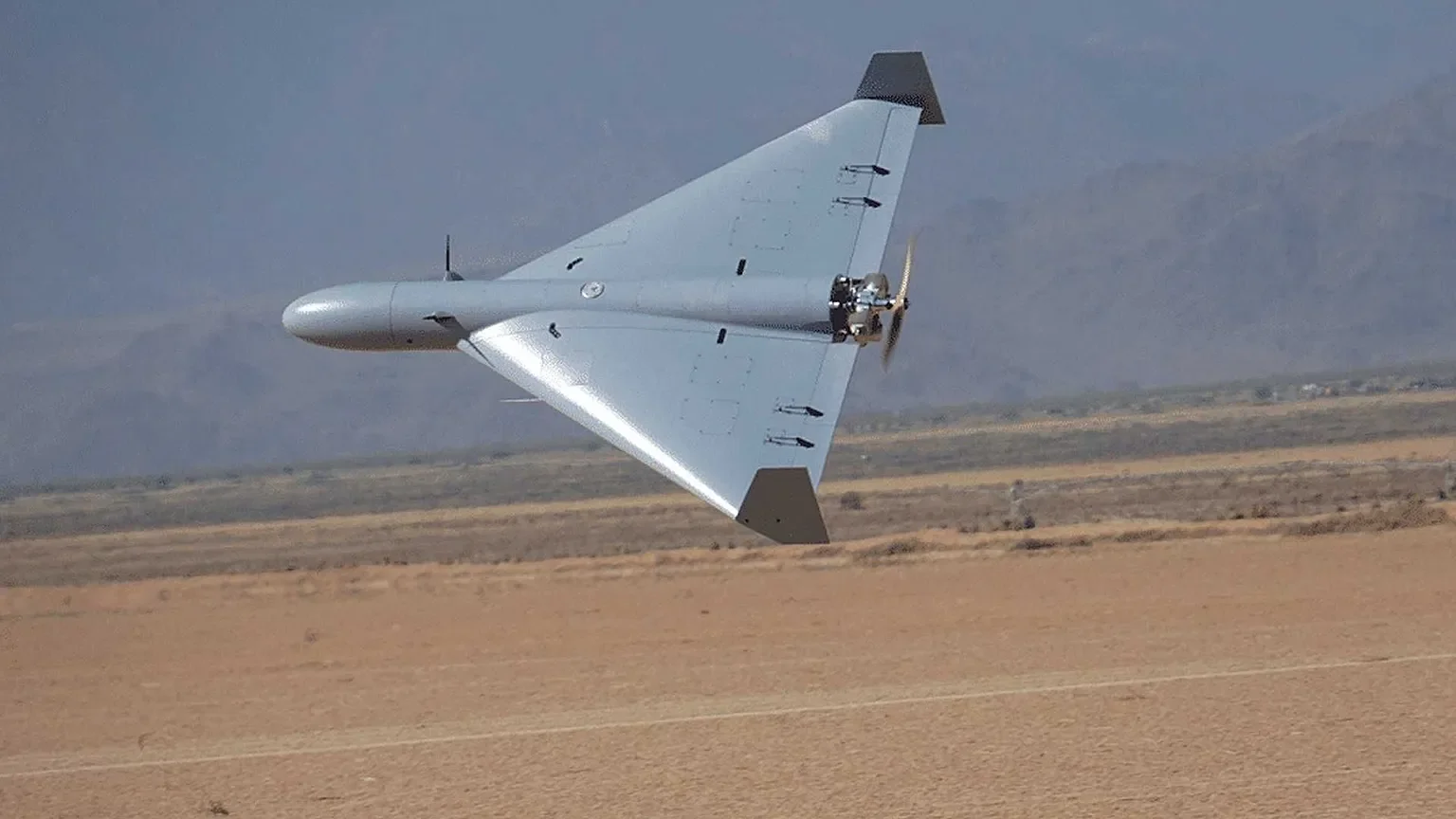 A military drone flying low over a flat desert landscape with mountains in the background.