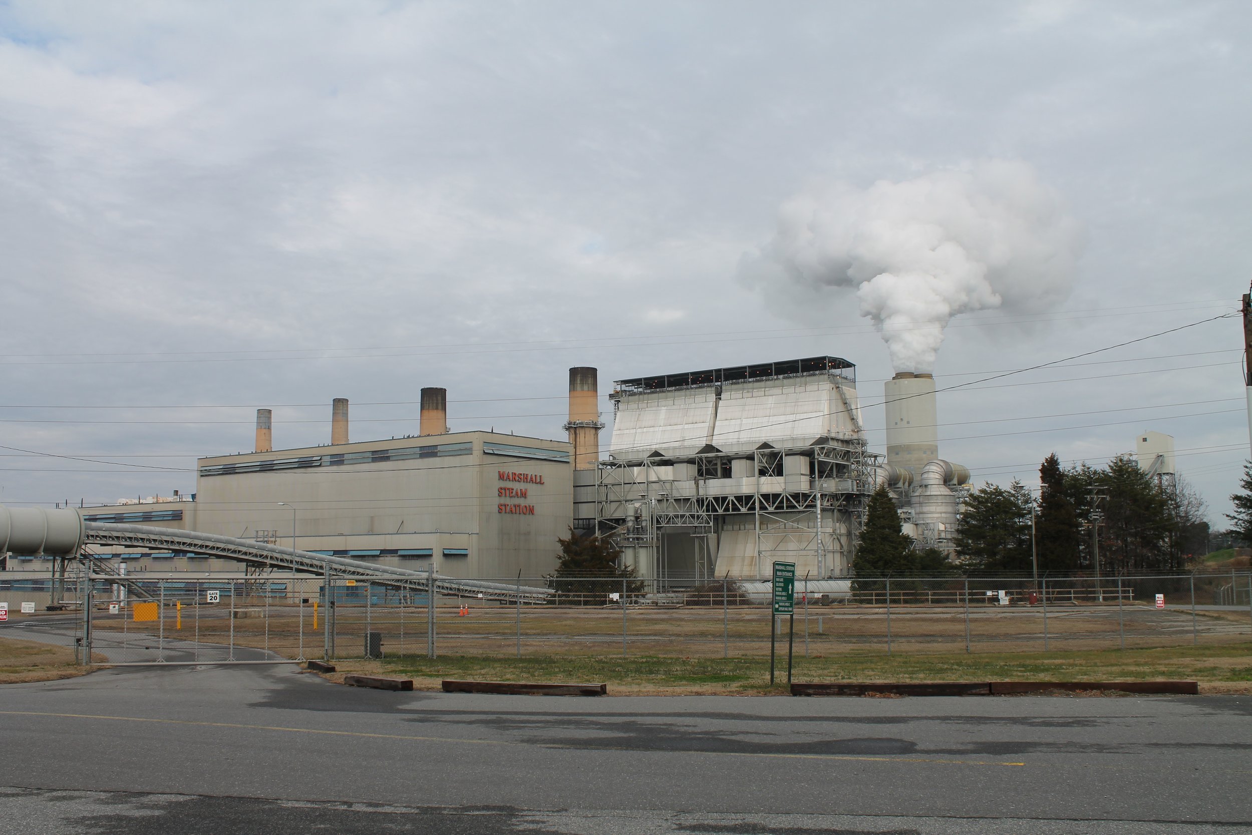 A power plant with several smokestacks emitting white smoke, labeled 'Marshall Steam Station,' with a chain-link fence in the foreground and a cloudy sky above.