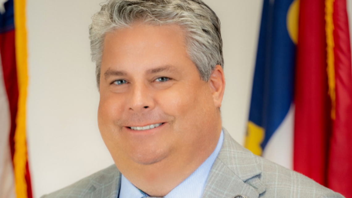 Portrait of a smiling man with gray hair wearing a light gray suit, standing in front of American flags.