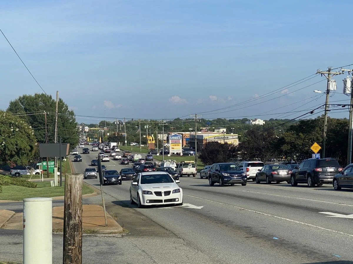 A busy traffic jam on a multi-lane road with cars, trucks, and a white sports car in the front. There are commercial stores and billboards visible in the background, along with utility poles and power lines, under a partly cloudy blue sky.