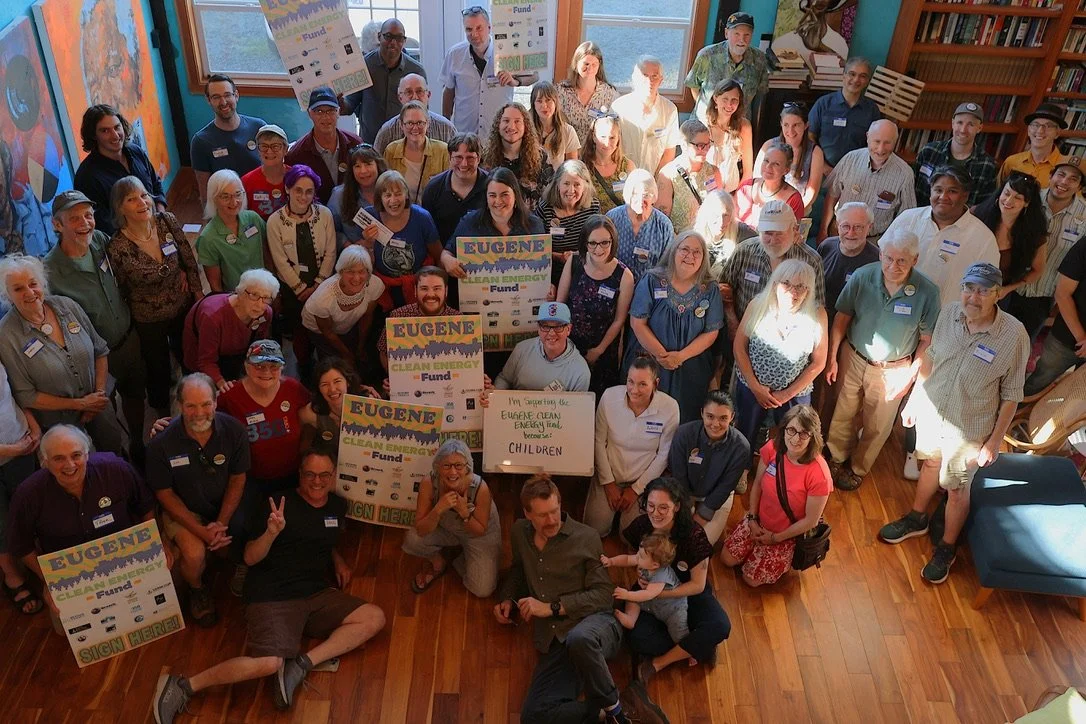 Group of people gathered indoors, holding signs that say 'Eugene Clean Energy Fund' and a whiteboard that reads 'I'm supporting the Eugene Clean Energy Trail for Children' at a community event.