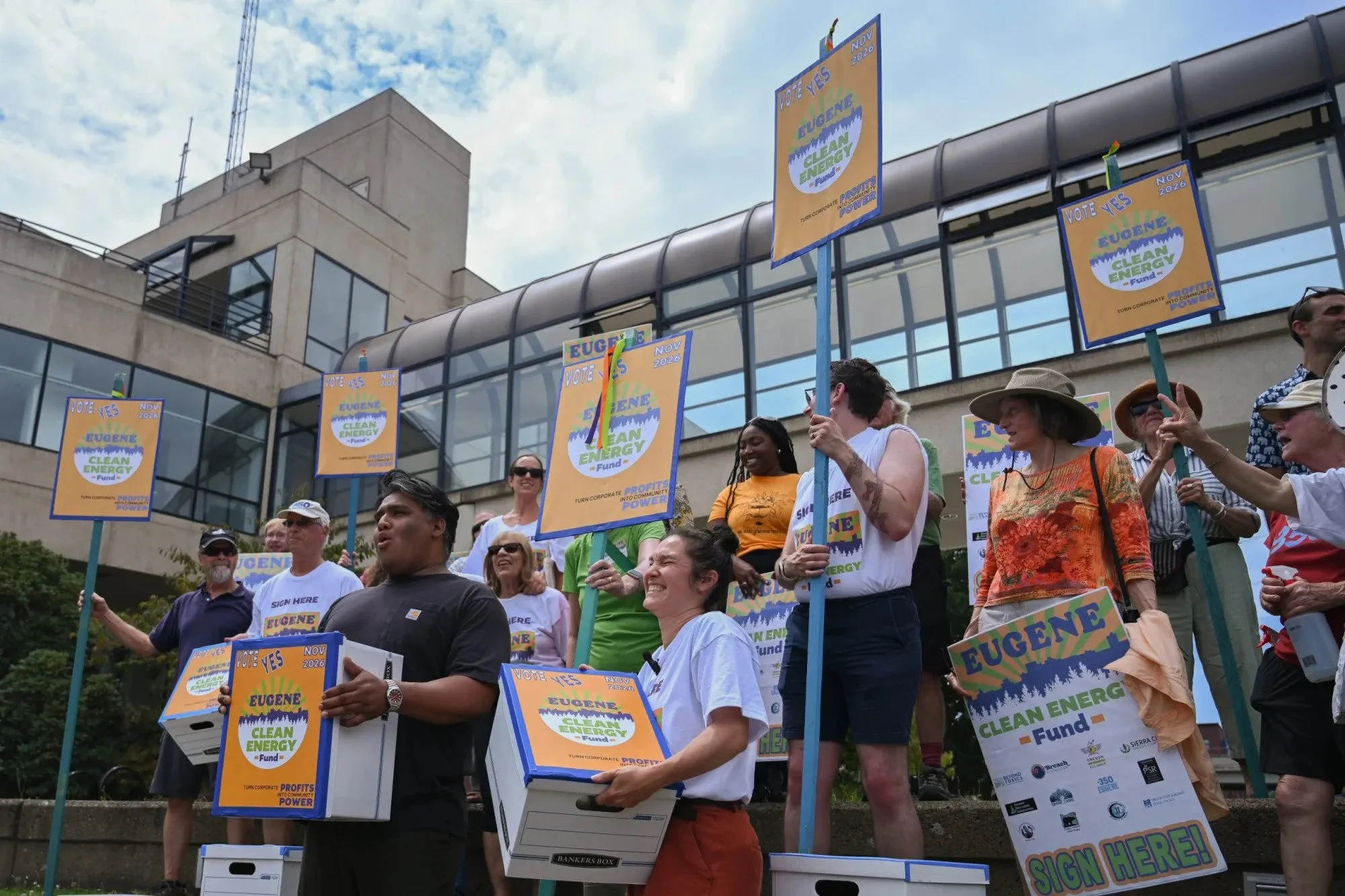 A group of people at a protest or rally holding signs that read "Vote Yes Eugene Clean Energy Fund" with a sun and mountain design. The signs indicate support for a November 2026 election. There is a modern building with large windows behind them and the sky is partly cloudy.