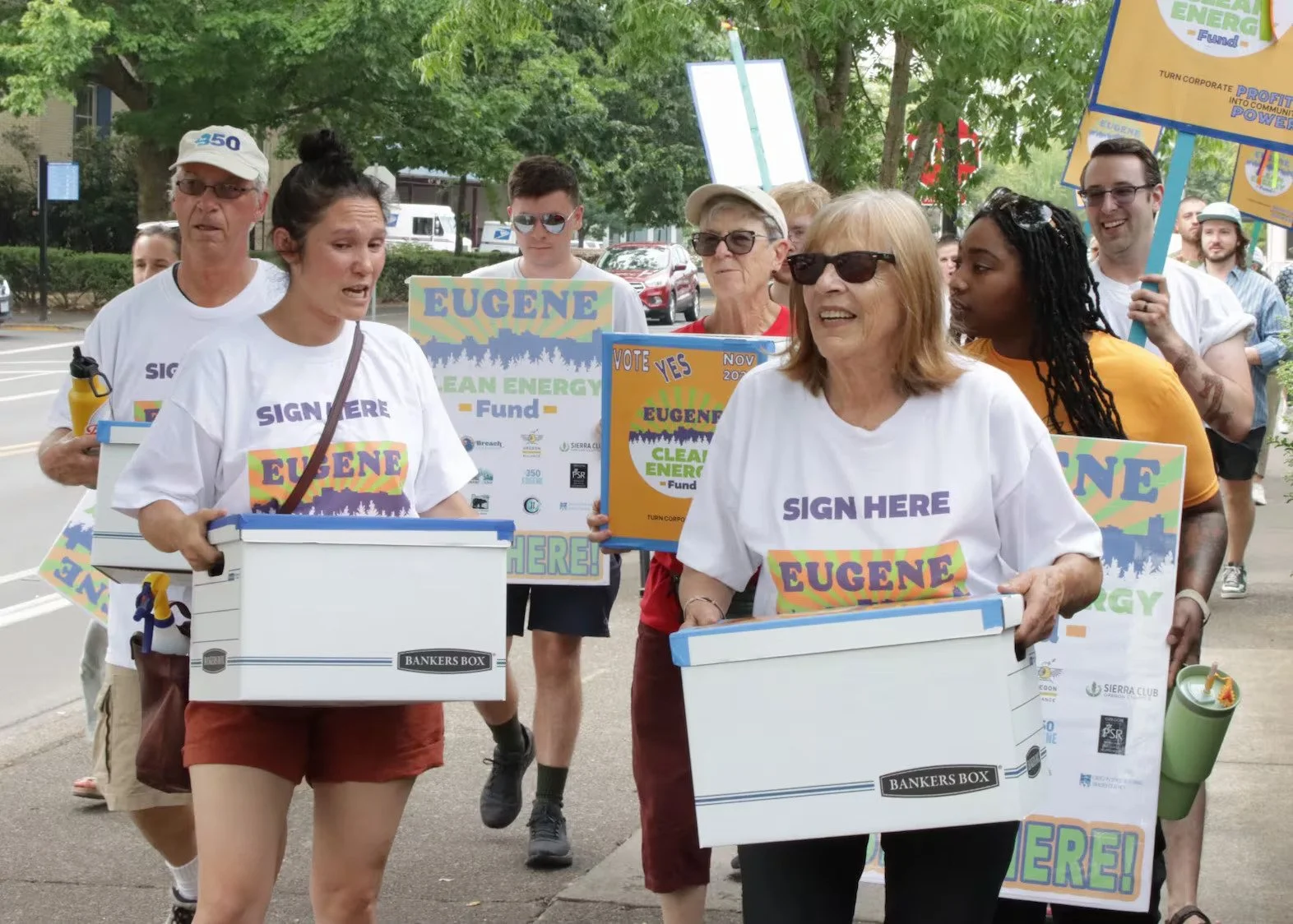 Group of people participating in a community event for clean energy, holding signs and donation boxes, with some wearing shirts that say "SIGN HERE EUGENE".
