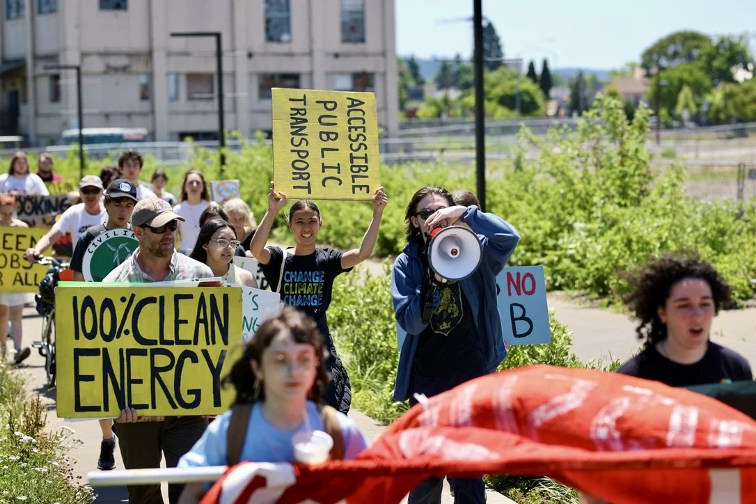 People participating in an outdoor climate protest march, holding signs with messages like "100% Clean Energy" and "Accessible Public Transport."