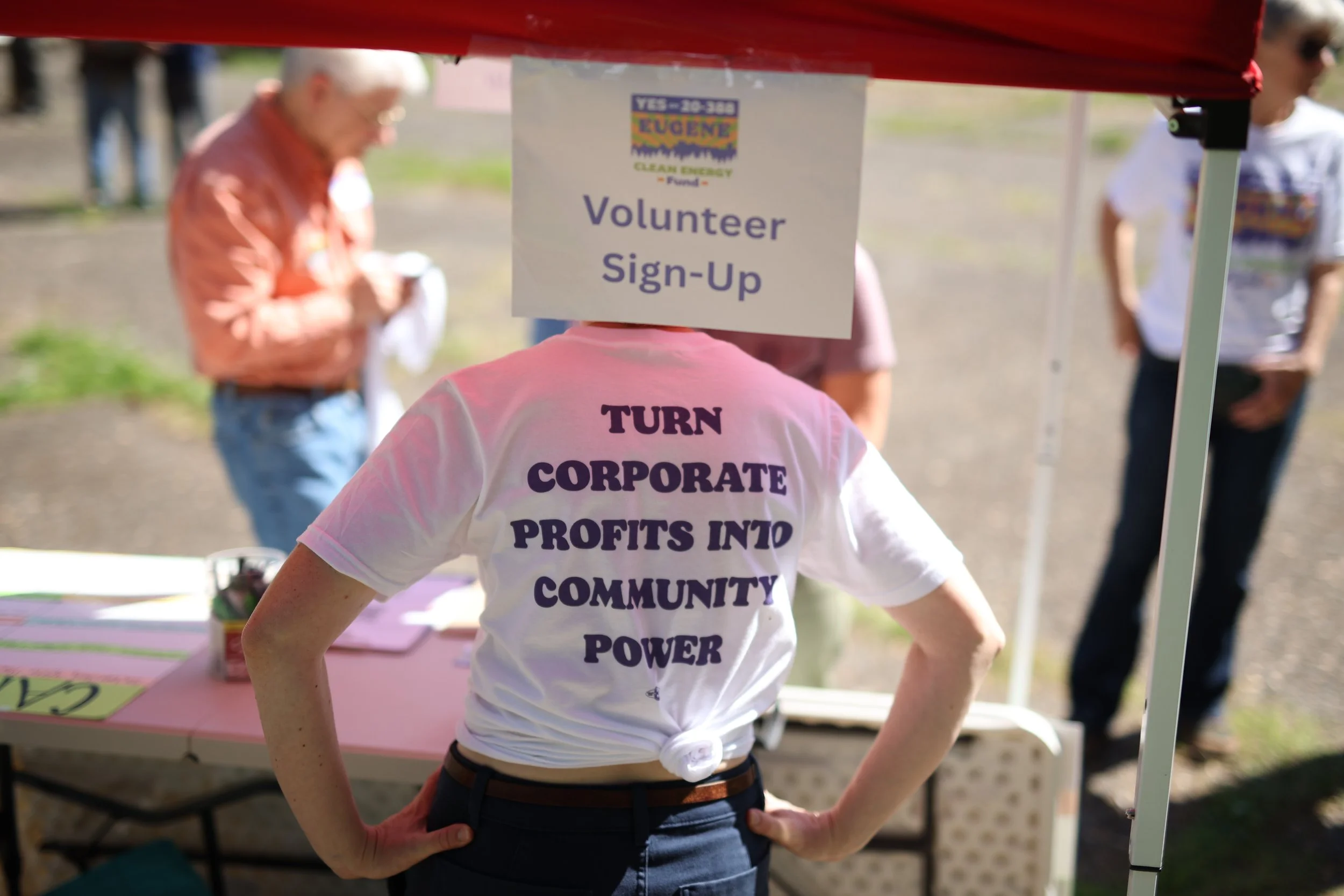 Person wearing a white t-shirt with purple text reading 'TURN CORPORATE PROFITS INTO COMMUNITY POWER' standing at a booth with a sign labeled 'Volunteer Sign-Up' at an outdoor event.
