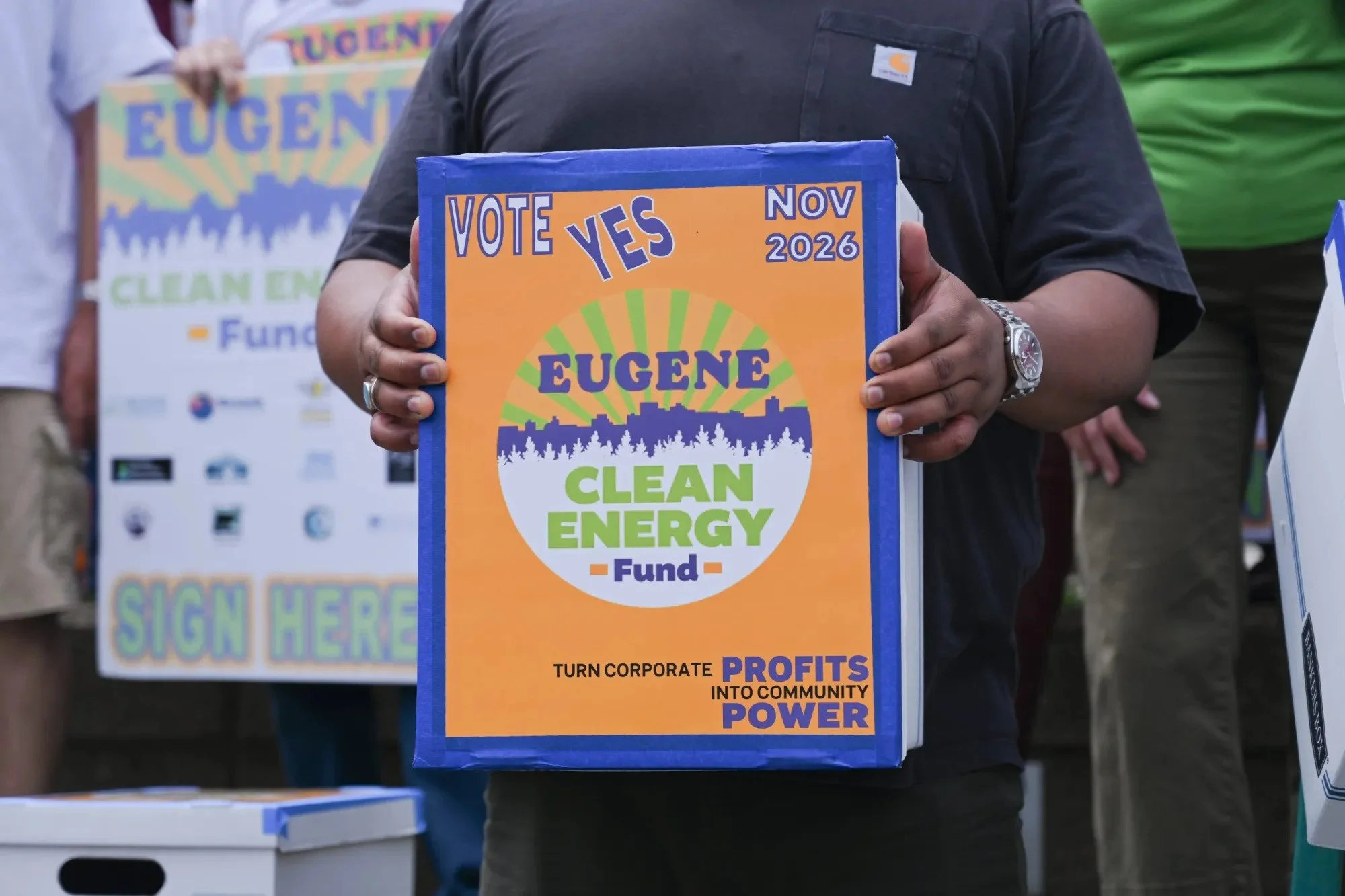 Person holding blue and orange sign promoting clean energy with the words 'VOTE YES' and 'EUGENE' on it. Other people are visible in the background at an outdoor event.