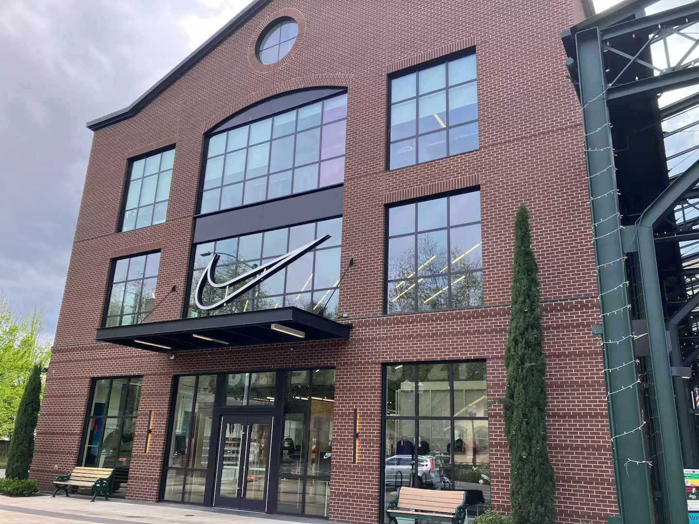 Exterior of a brick Nike store with large glass windows, a Nike swoosh logo, and a glass staircase on the side. There are trees and benches outside.