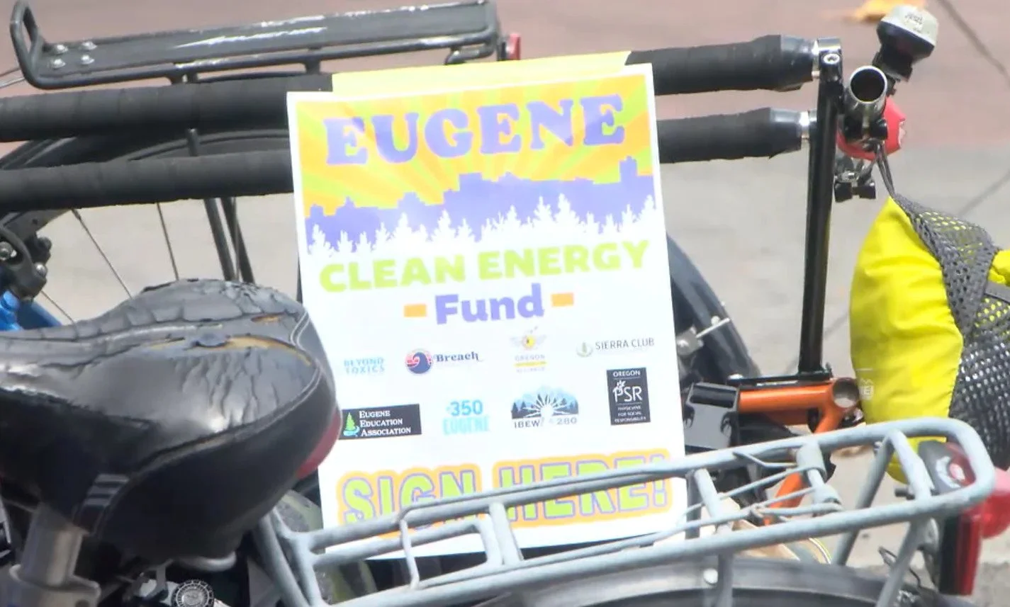 A bicycle with a sign attached to it displays the name 'Eugene' and promotes a 'Clean Energy Fund' with various sponsor logos, including Eugene Education Association and Oregon PSR.
