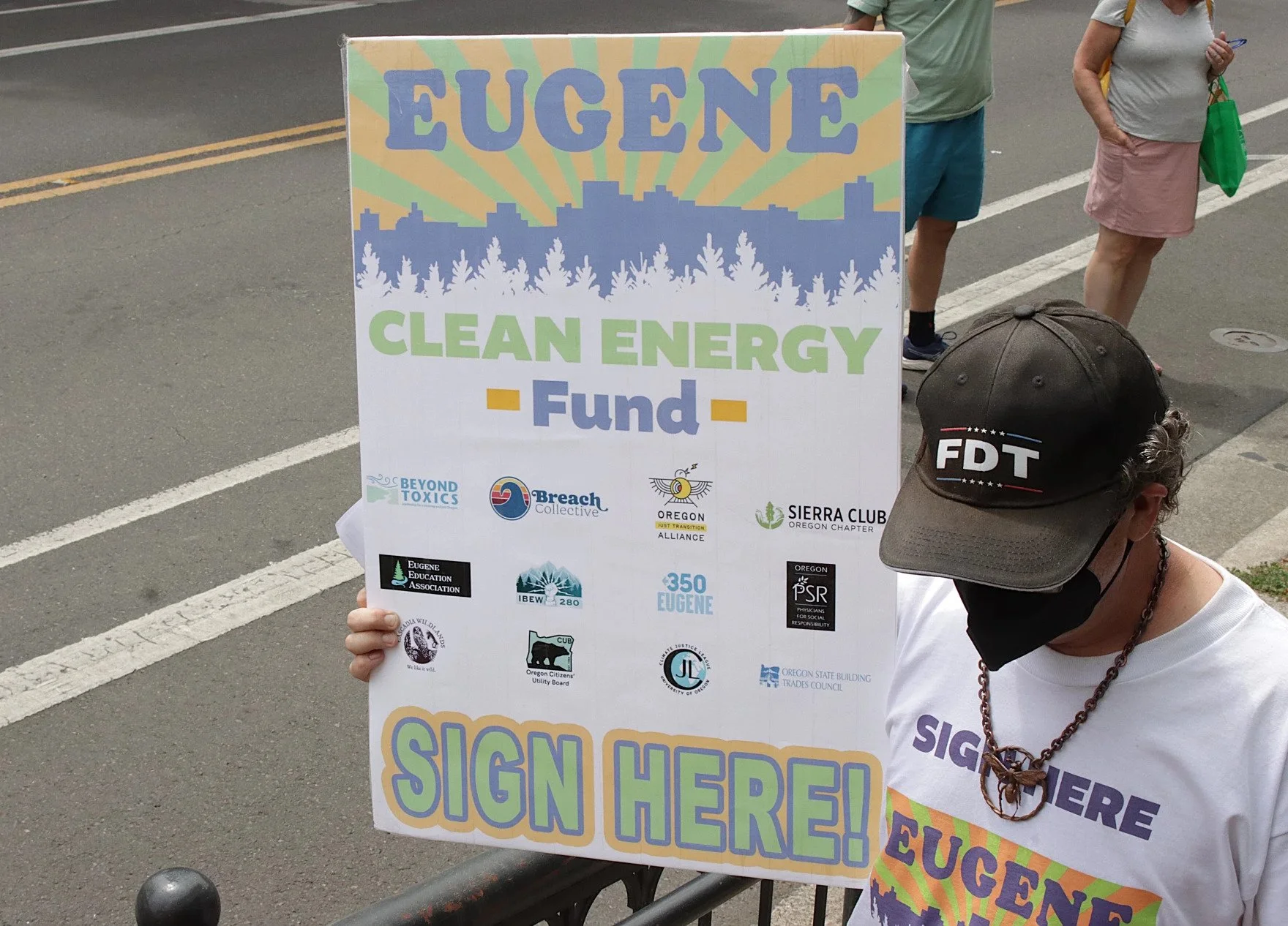 A person holding a sign that reads 'Eugene Clean Energy Fund' with logos of various organizations at a protest or rally on an urban street. The sign has a colorful design with a city skyline and trees.