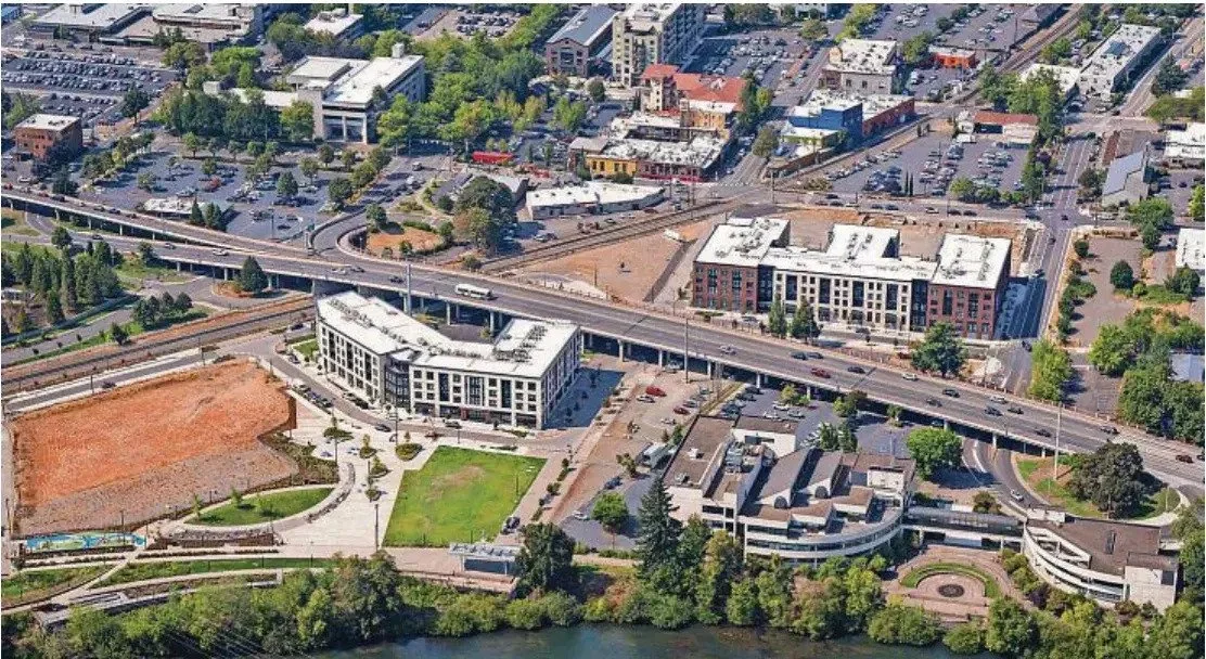 An aerial view of an urban area with a highway running through it, surrounding buildings and parking lots, a park with trees and a green lawn, and a body of water with a walking path.