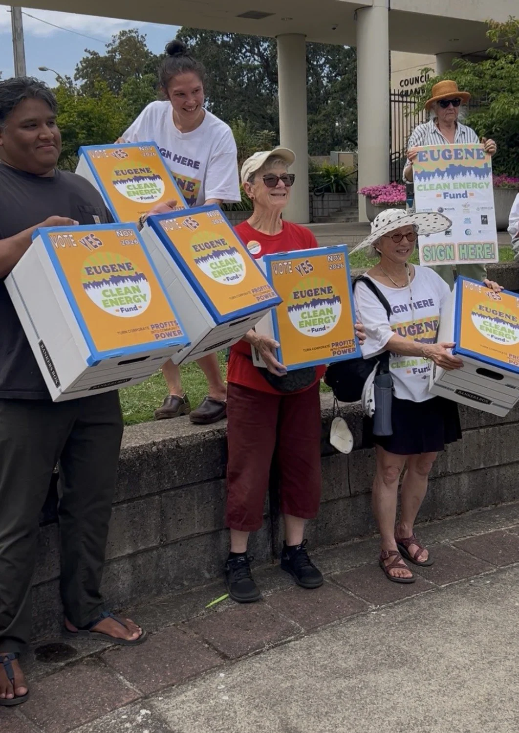 A group of diverse people holding boxes and signs supporting the Eugene Clean Energy Fund, participating in a community event or campaign.