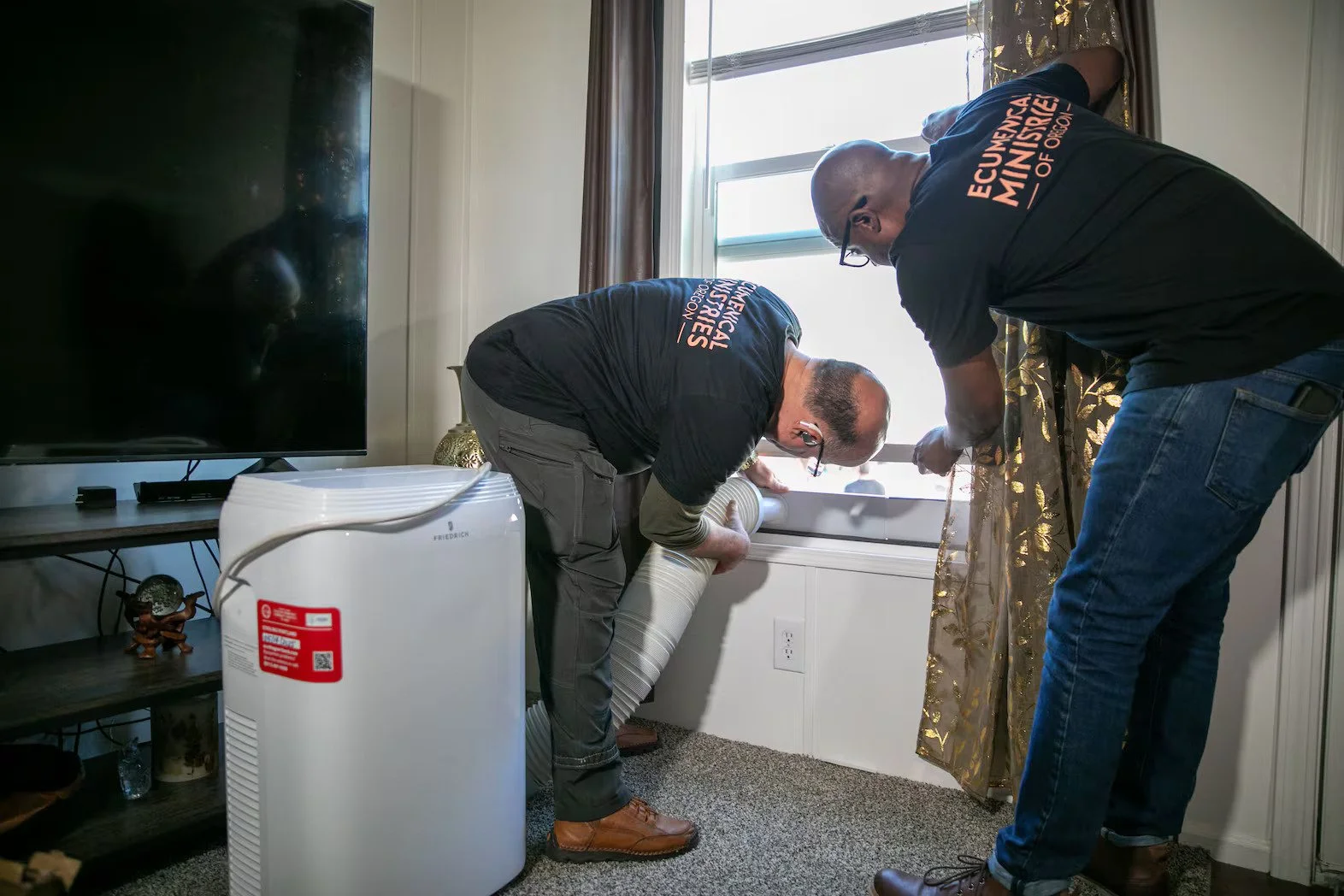 Two technicians installing or repairing a window air conditioning unit in a living room, one is bending down adjusting the unit while the other is holding a tool near the window.