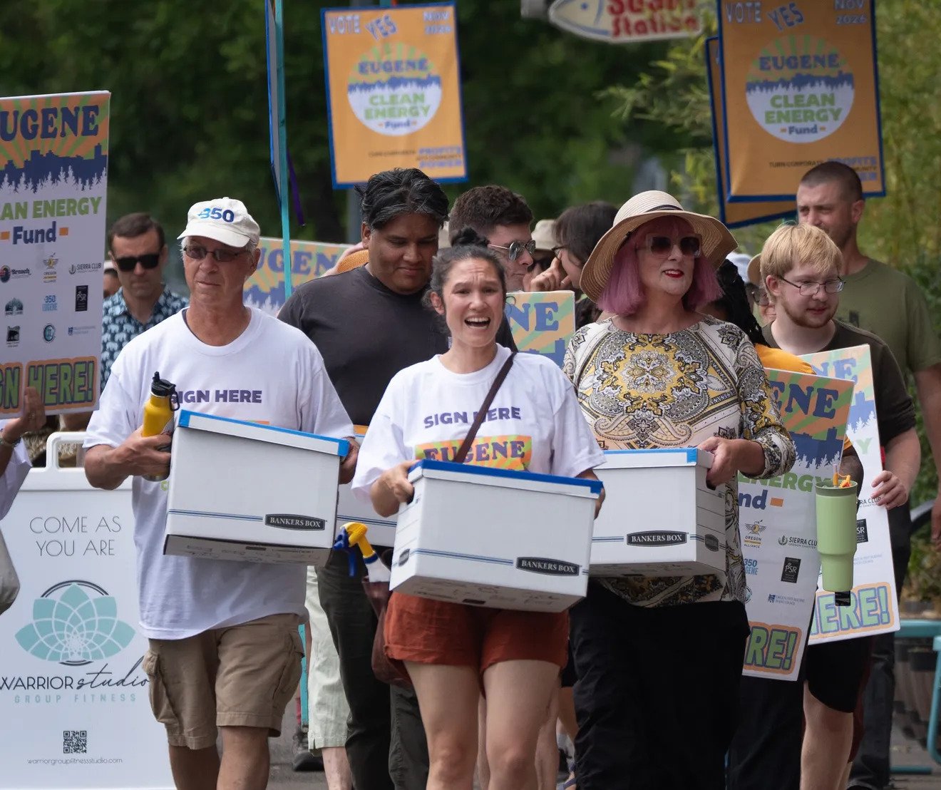 People participating in an outdoor rally or march holding signs and boxes, with banners promoting Eugene Clean Energy Fund in the background.