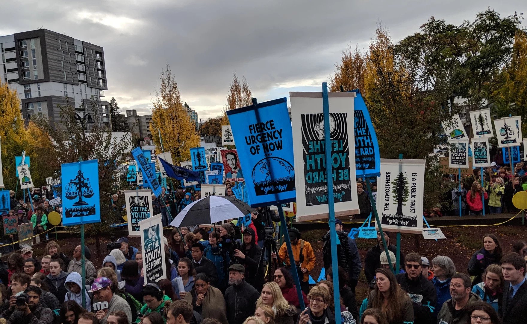 A large crowd of young people and adults gathered outdoors for a protest or rally, holding signs and banners with messages about climate change and youth activism, with trees and buildings in the background.