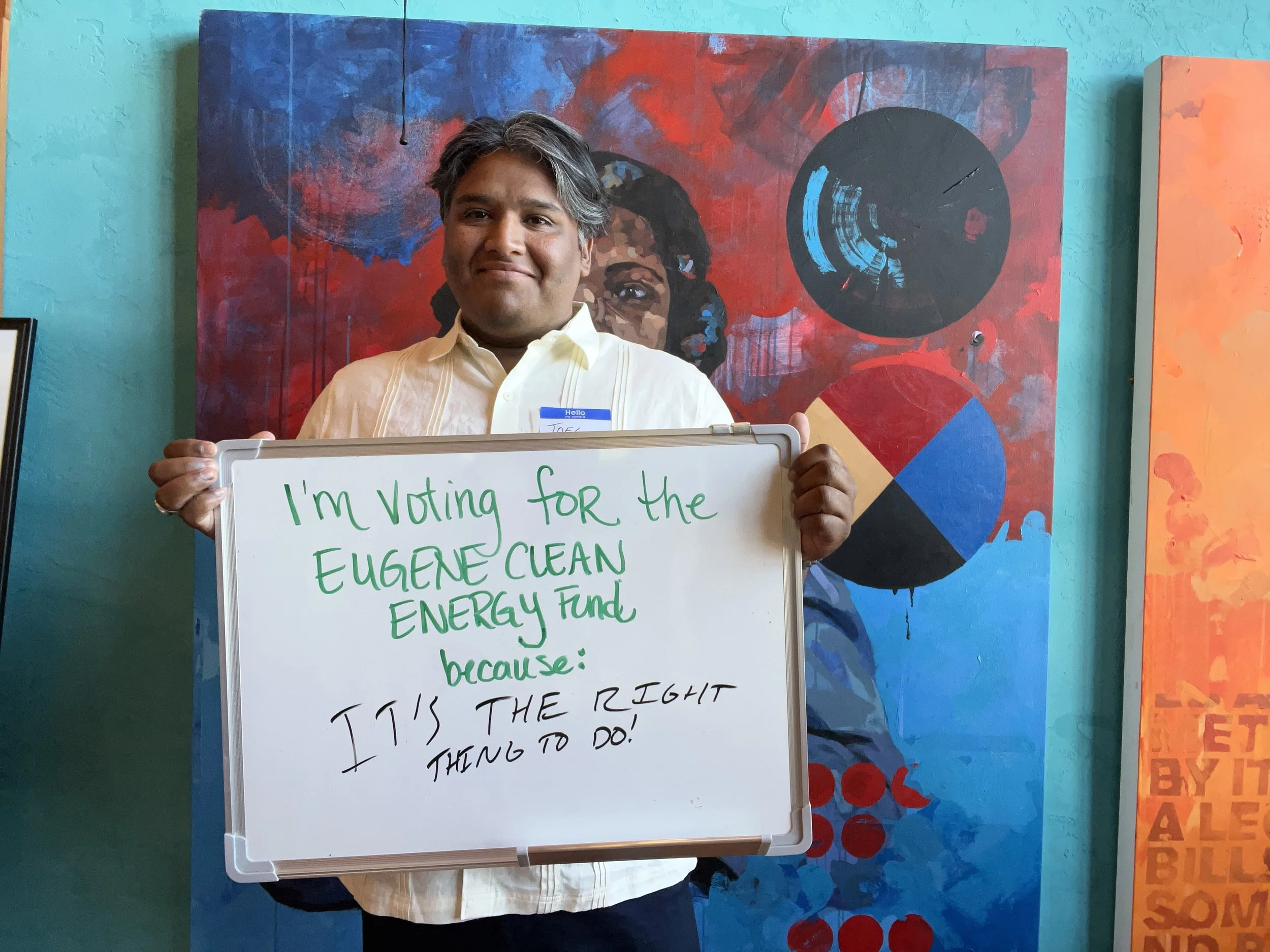 A man with gray hair and a light-colored shirt is holding a whiteboard with writing on it. There is an abstract, colorful painting in the background.