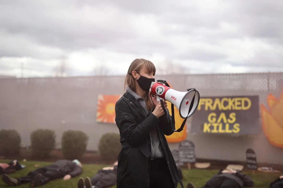 A woman wearing a black face mask holding a megaphone at a protest or climate rally, with people lying on the ground and a sign in the background that reads 'FRACKED GAS KILLS'.