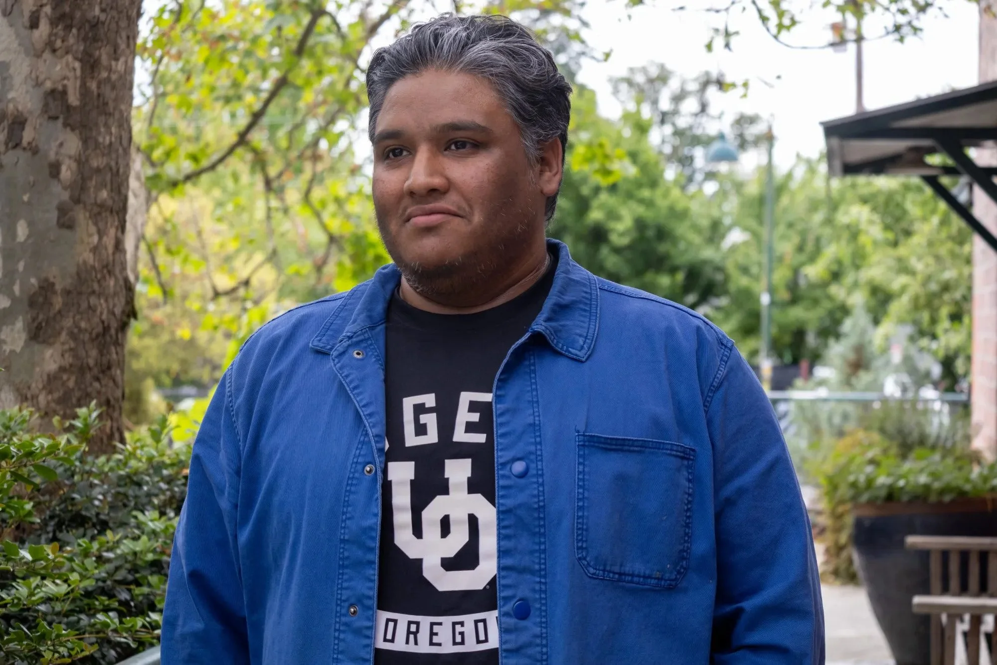 A man with dark hair and medium skin tone standing outdoors near trees and greenery, wearing a blue jacket over a black T-shirt with white lettering.