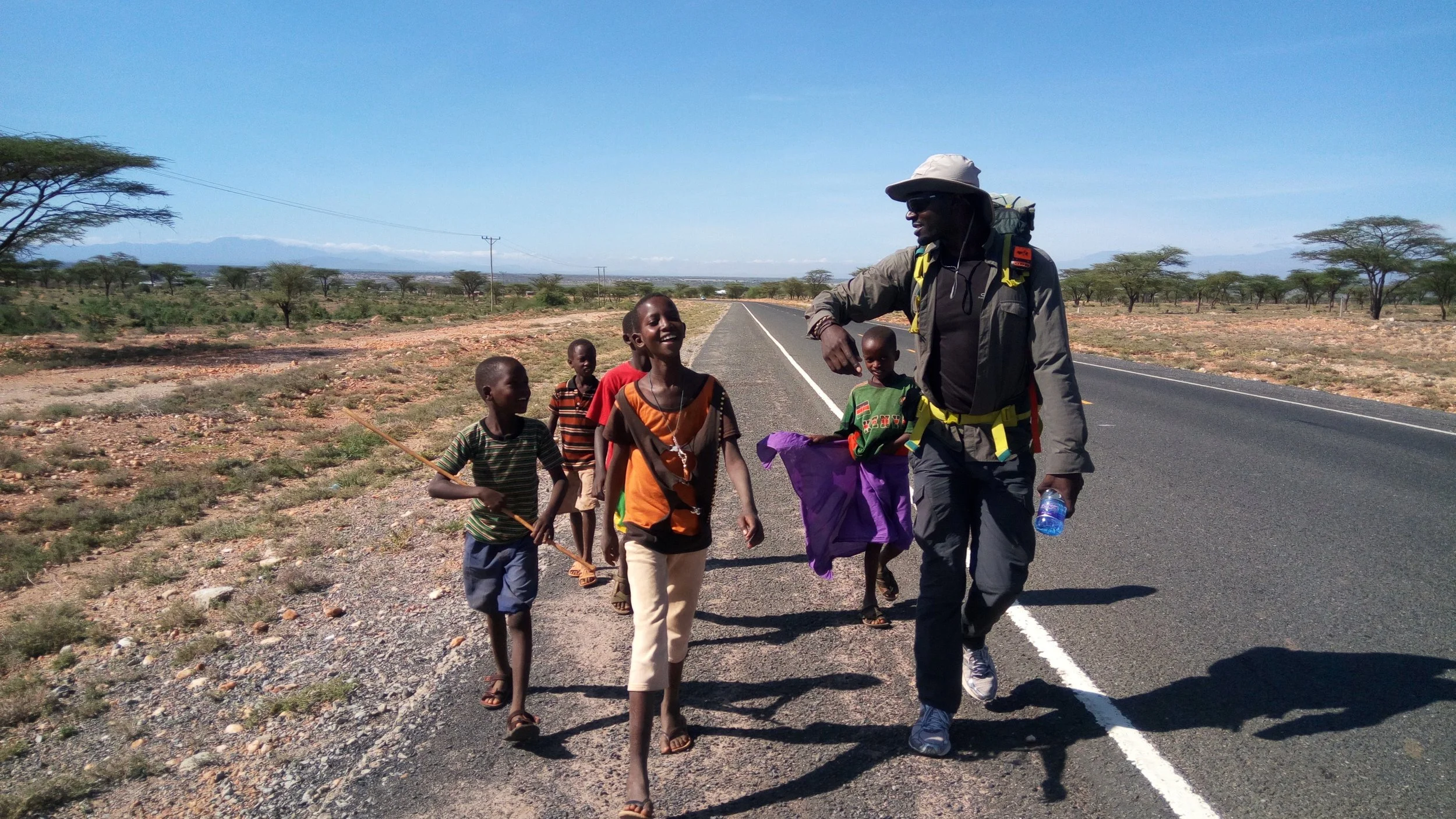 A man with a backpack and hat walking along a deserted road in a rural area, surrounded by children smiling and walking with him. The landscape is dry with sparse trees under a clear blue sky.