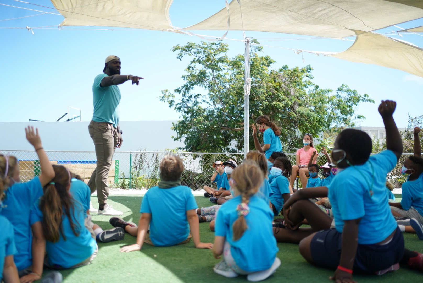 A man standing on a grassy area speaking and gesturing to a group of children seated on the ground, many of whom have raised their hands. The children are wearing face masks, and the setting appears to be outdoors under shade sails with trees and a fence in the background.