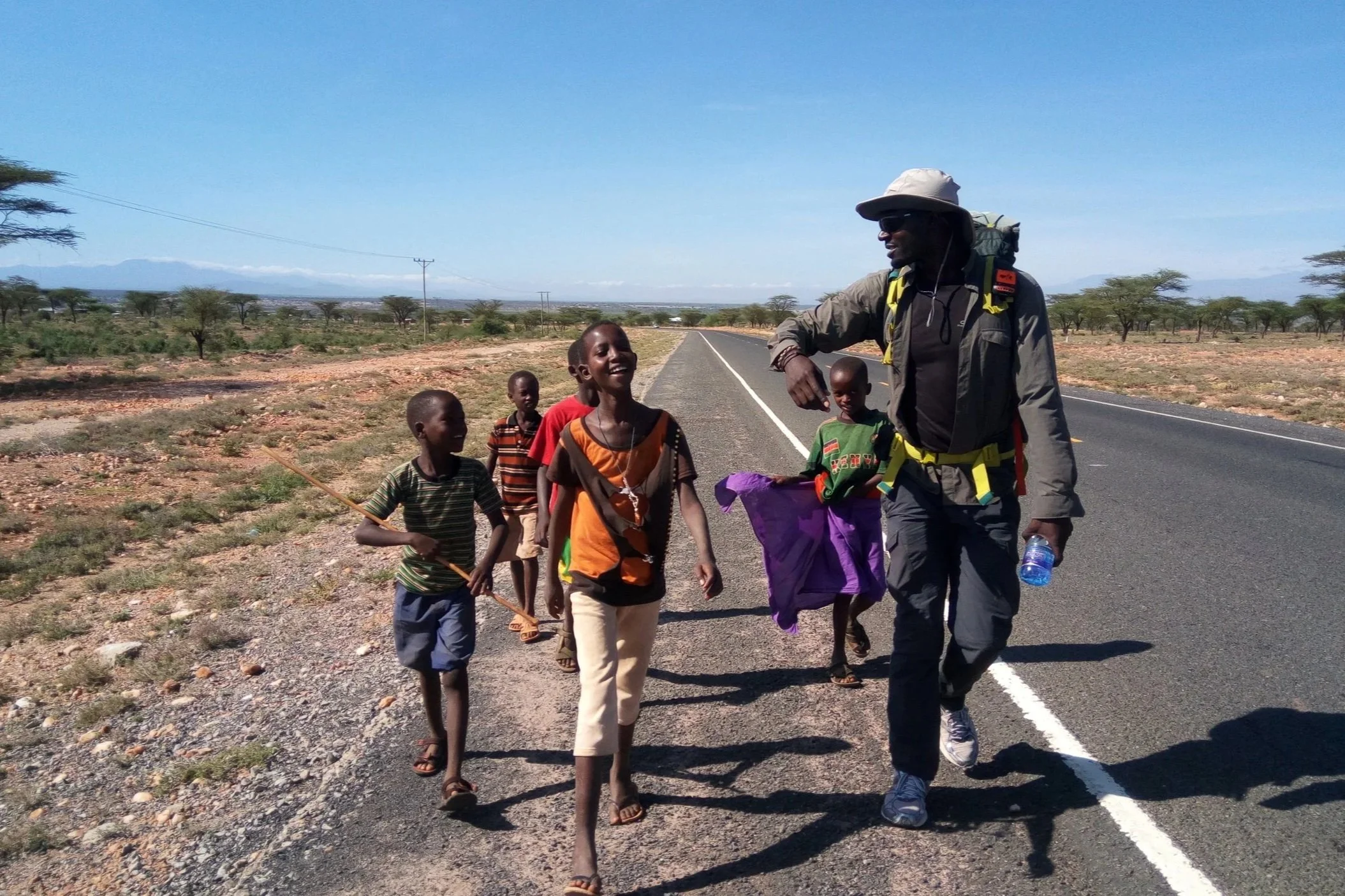 A man with a backpack and hat walking on the side of a rural highway, chatting with five smiling children in colorful clothes in a dry, flat landscape with sparse trees and mountains in the distance.