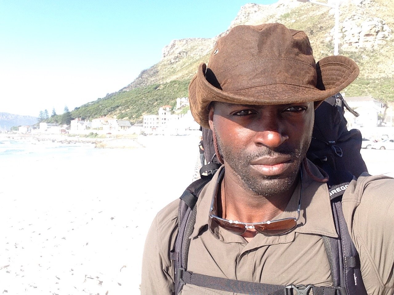 A man wearing a brown wide-brimmed hat, sunglasses hanging from his shirt collar, and a backpack, standing on a bright beach with cliffs and houses in the background.