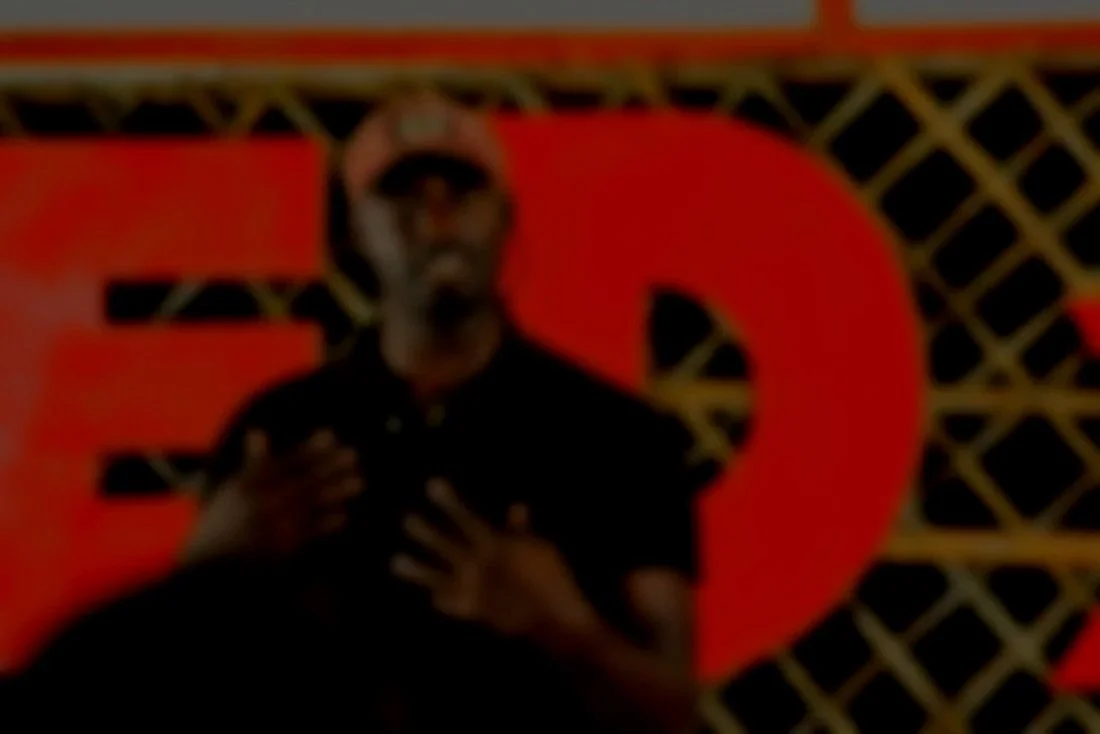 A person wearing sunglasses, a hat, and black clothing appears to be taking a selfie in front of a background with a large red letter 'E' and a black and yellow geometric pattern.