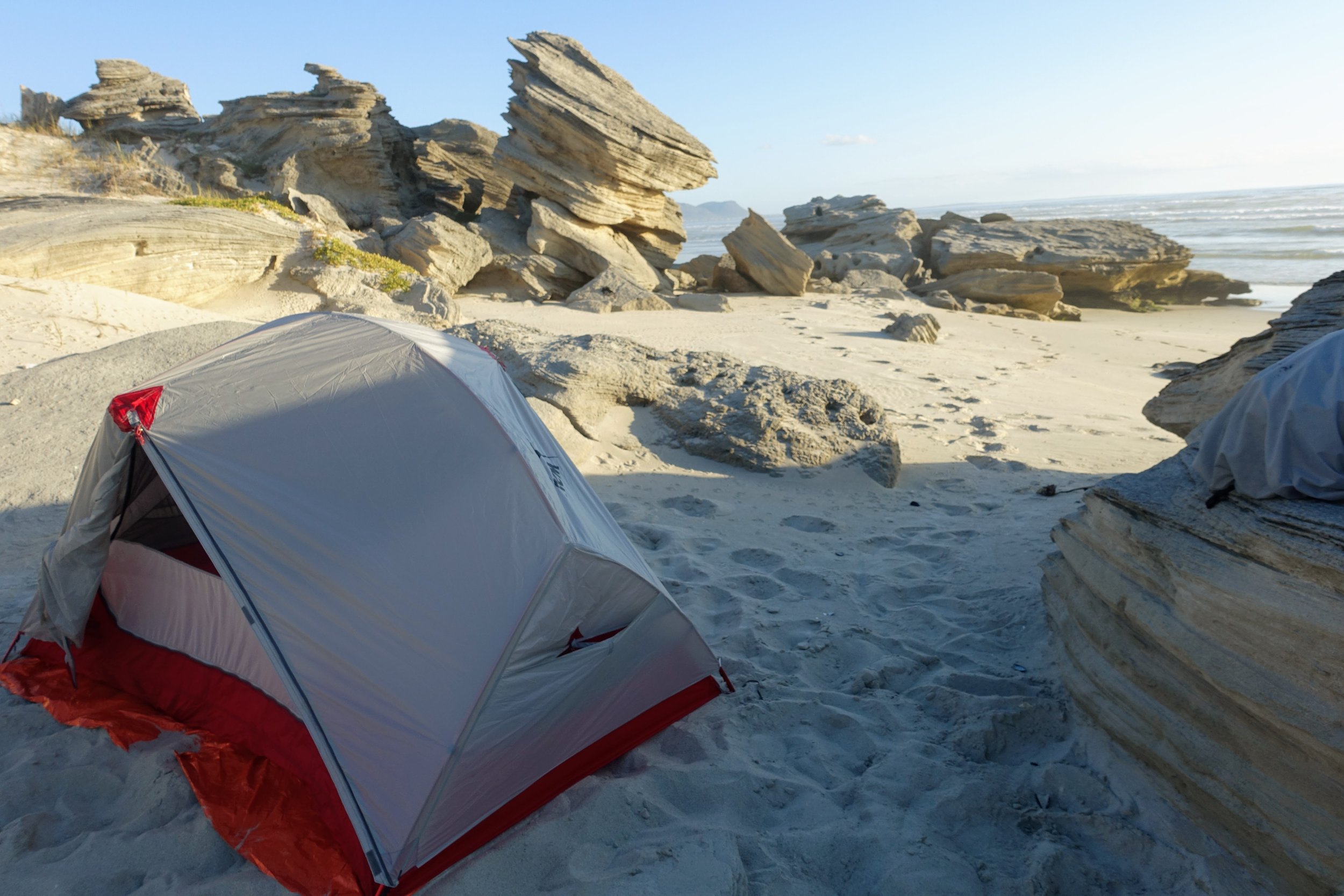 A camping tent set up on a sandy beach with large rock formations in the background and the ocean in the distance.