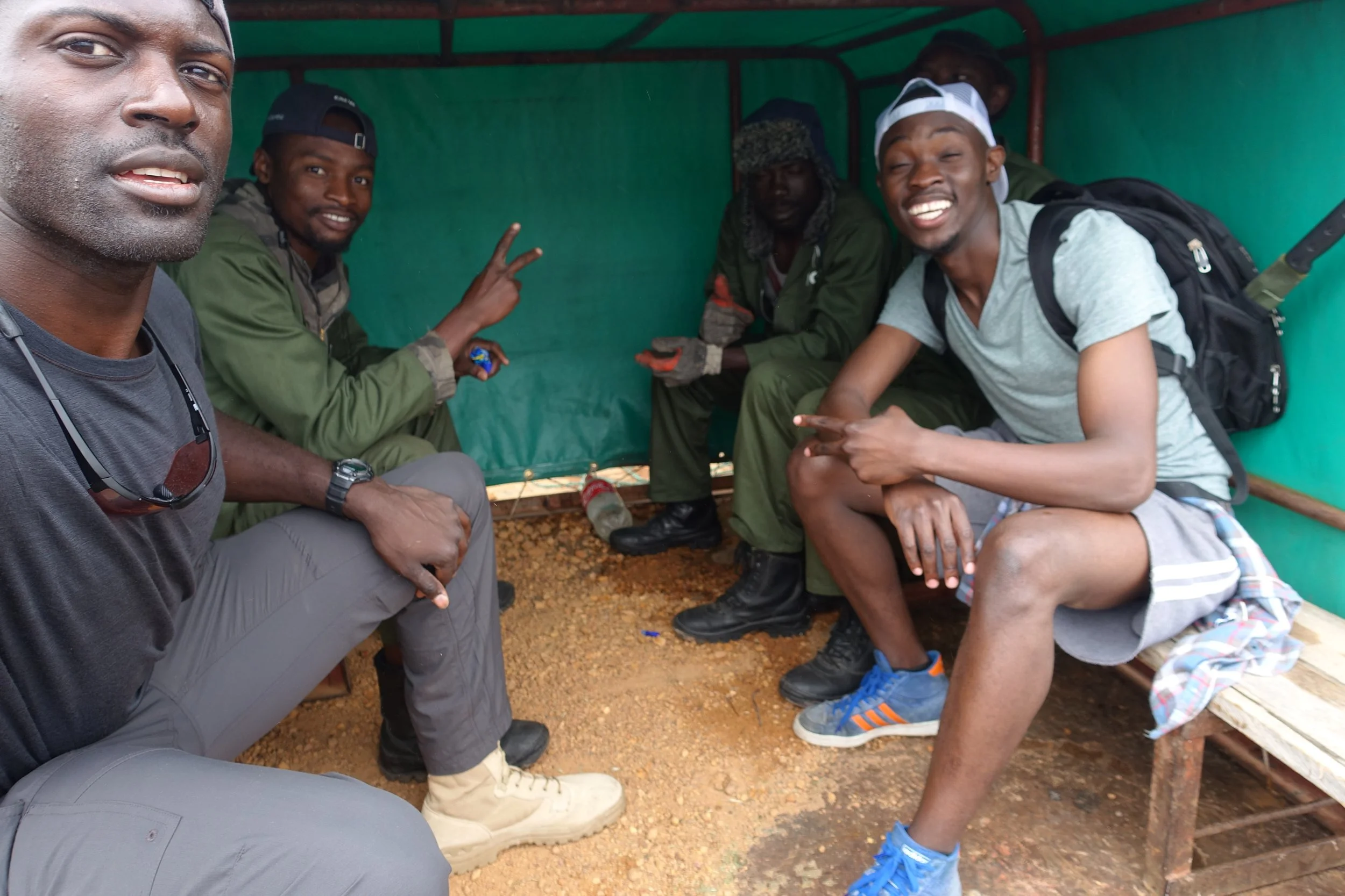 Five young men sitting inside a makeshift shelter with a green tarpaulin roof and walls, smiling and posing for the photo. One is taking the selfie, while others are making peace signs or pointing at the camera.