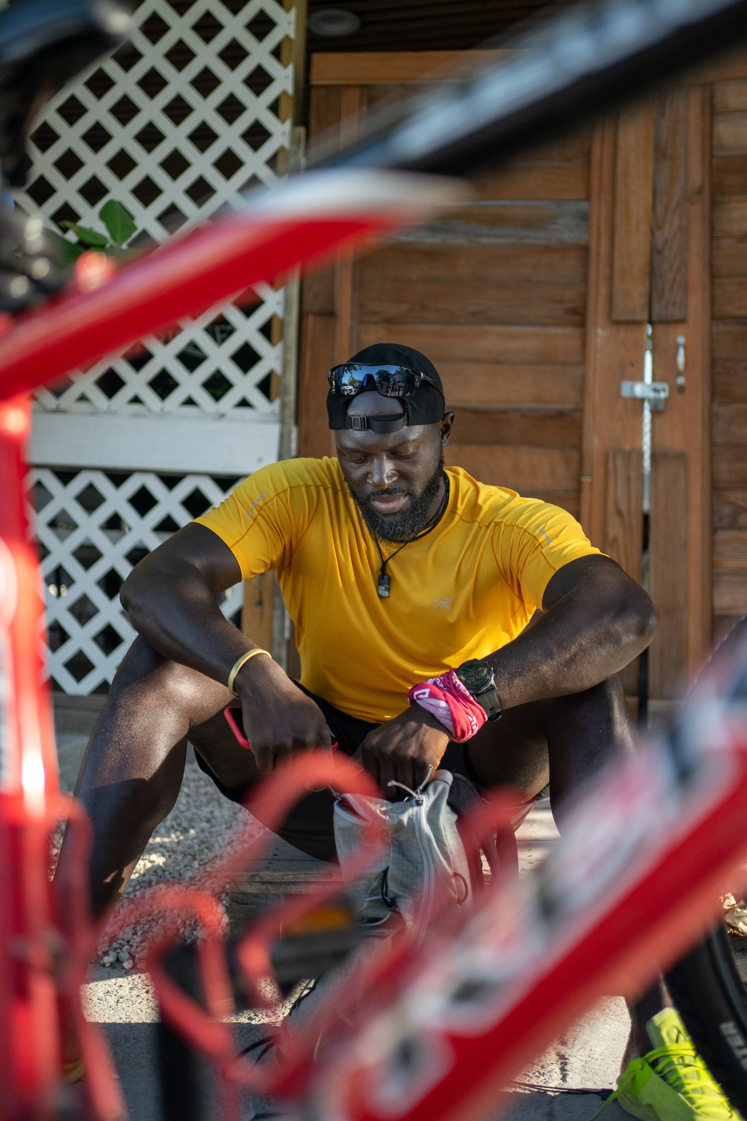 A man wearing a yellow athletic shirt, a black cap, sunglasses on his head, and a red bandana on his wrist, sits on the ground, appears to be preparing for biking, with a red bicycle frame partially visible in the foreground and a wooden shed in the background.