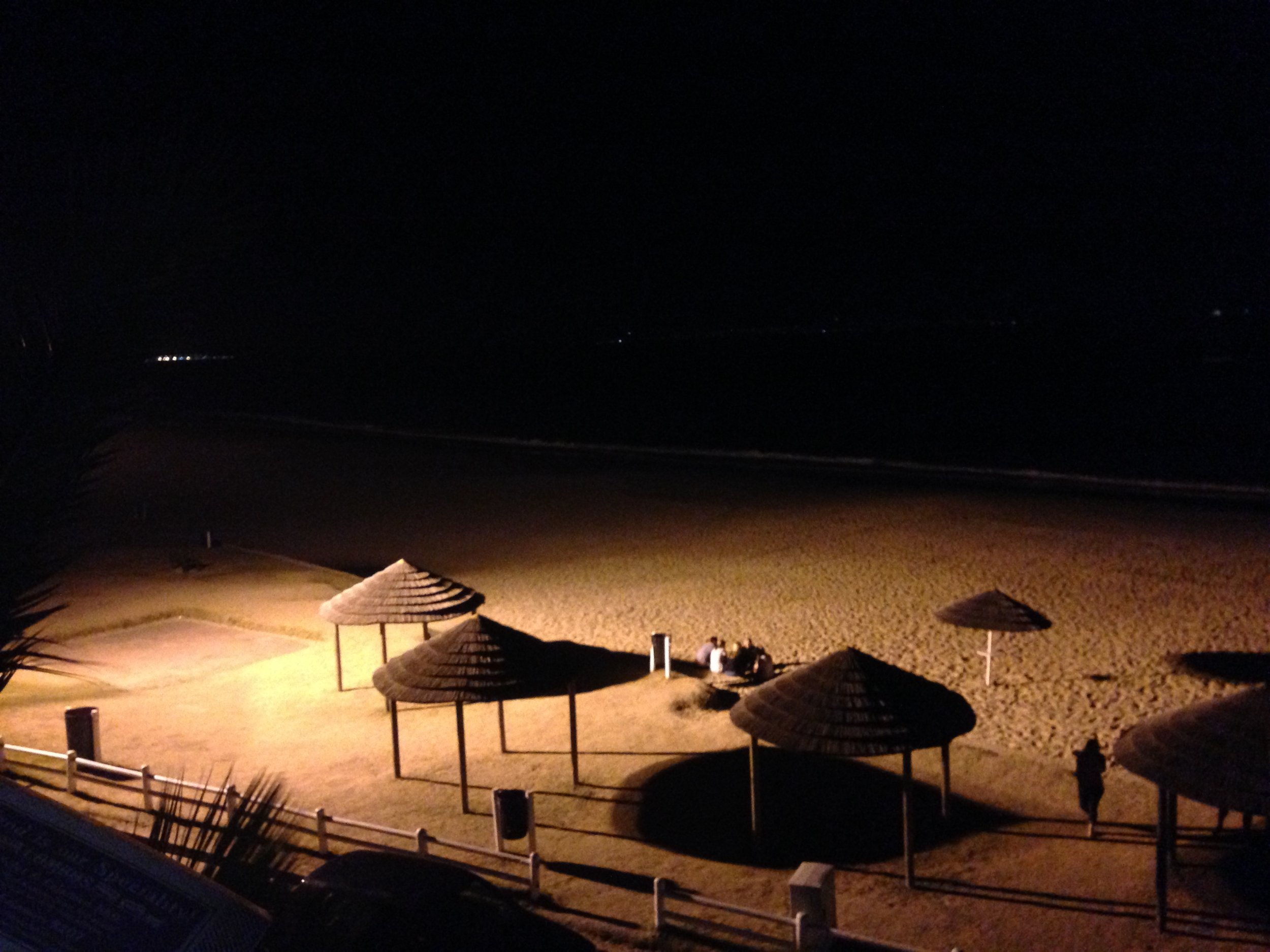 Nighttime view of a beach with several thatched umbrellas, some people sitting on benches, and a person walking along the sand, illuminated by artificial lights.