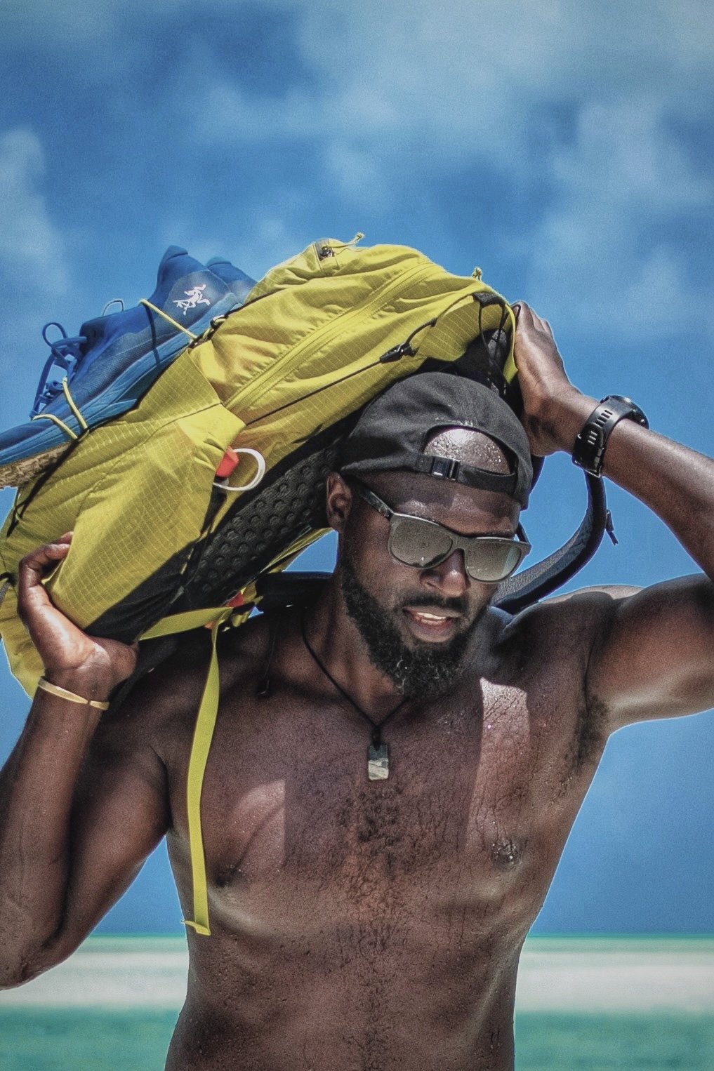 A shirtless man carrying a yellow backpack on his shoulder, wearing sunglasses and a cap, on a sunny beach with blue sky and ocean in the background.