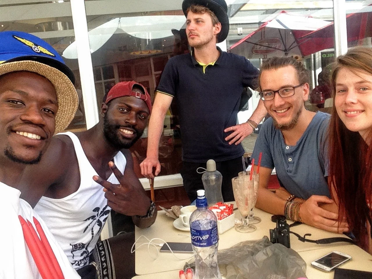 A group of five friends sitting at an outdoor cafe table, smiling at the camera. The table has drinks, water bottles, and a camera, with a man standing behind wearing a black hat and black shirt.