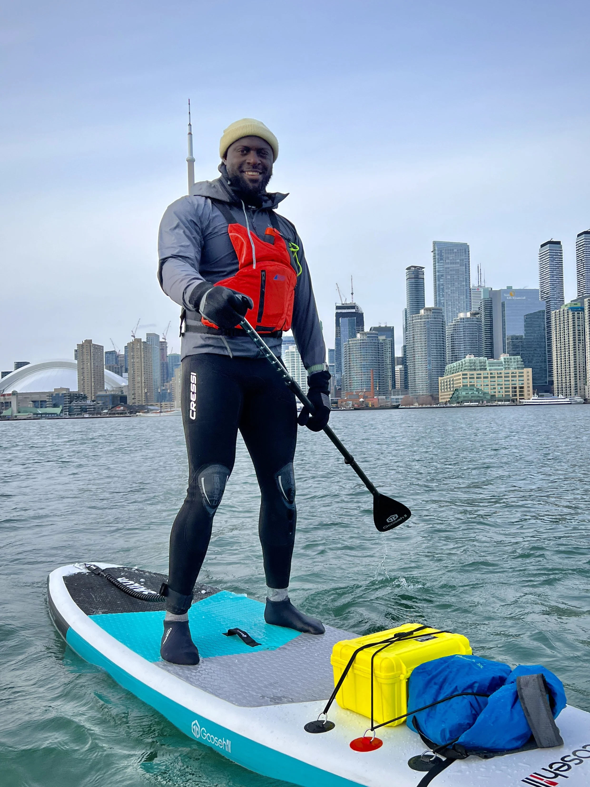 Man stand-up paddleboarding on water with city skyline behind him, wearing a red life jacket, black wetsuit, and beige beanie, holding a paddle.