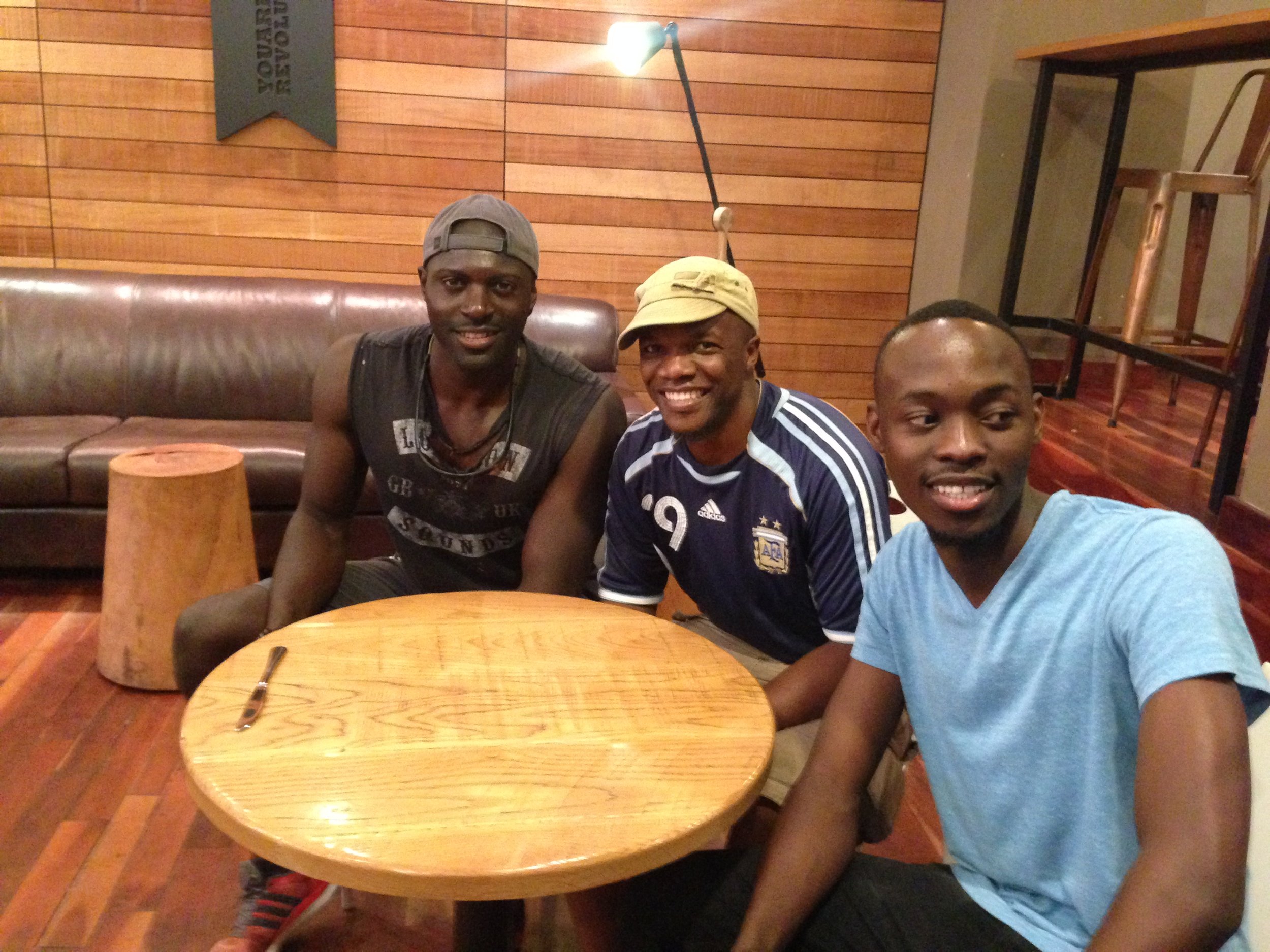 Three young men sitting at a wooden table in a cozy room with wooden wall paneling, smiling at the camera.