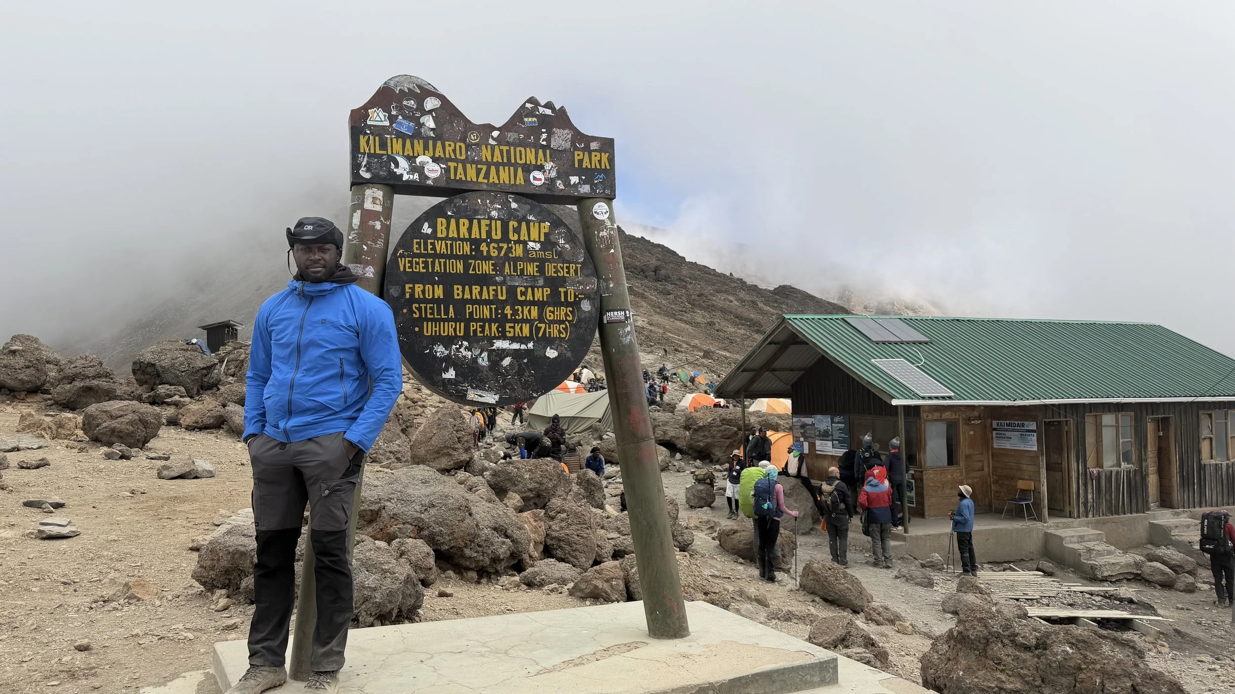 A man in a blue jacket and gray pants standing next to a sign at Kilimanjaro National Park in Tanzania, with a mountain and cloudy sky in the background, and a small wooden building with solar panels on the roof nearby.