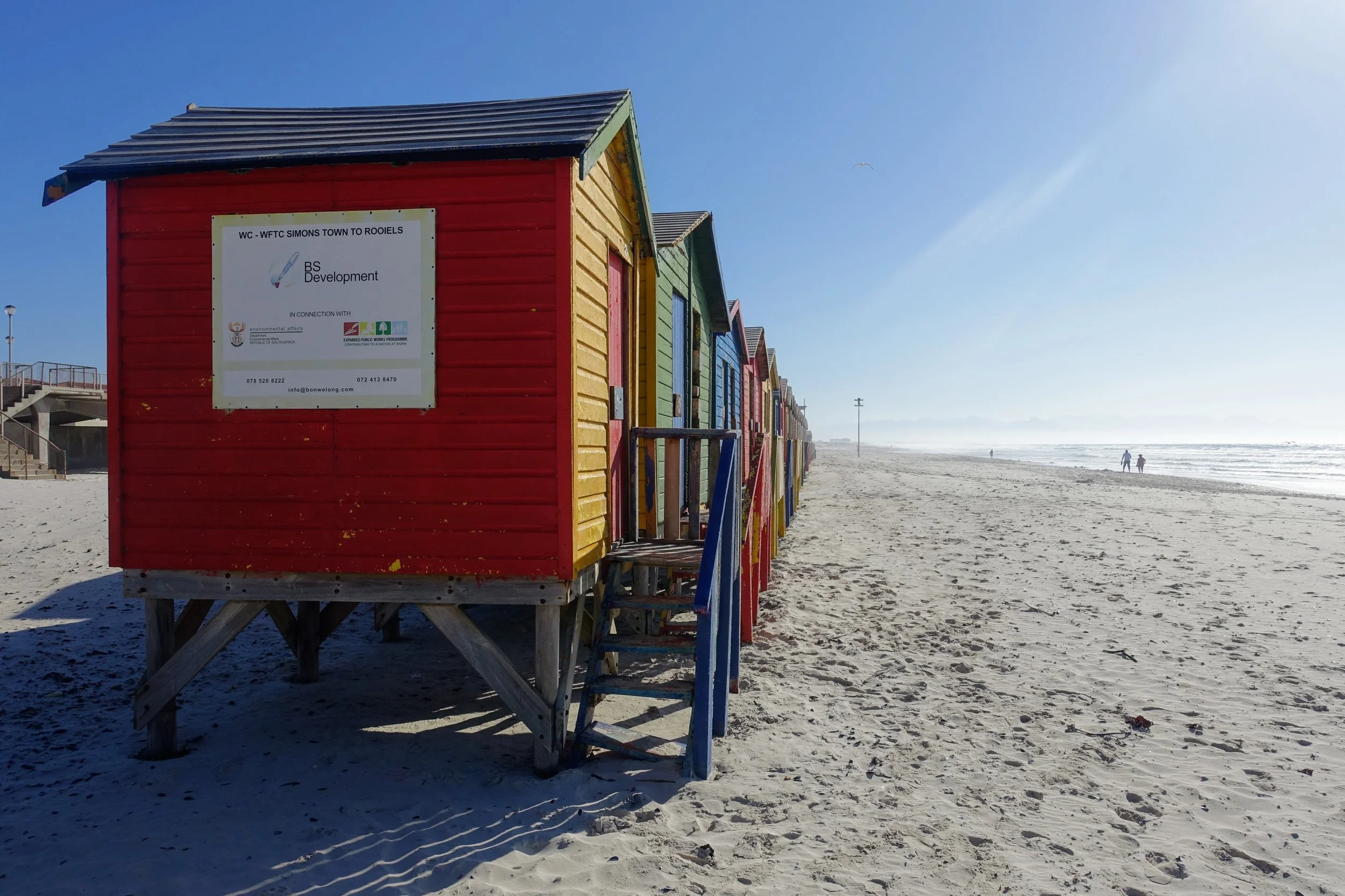 Colorful beach huts with a sign on the red hut in the foreground, set on a sandy beach under a clear blue sky with people walking along the shoreline in the distance.