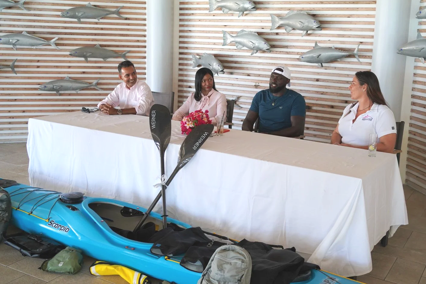 A panel of four people sitting at a table with a white tablecloth, in front of a wall decorated with inflatable fish and paddleboards, with water sports equipment like a kayak and paddles visible in the foreground.