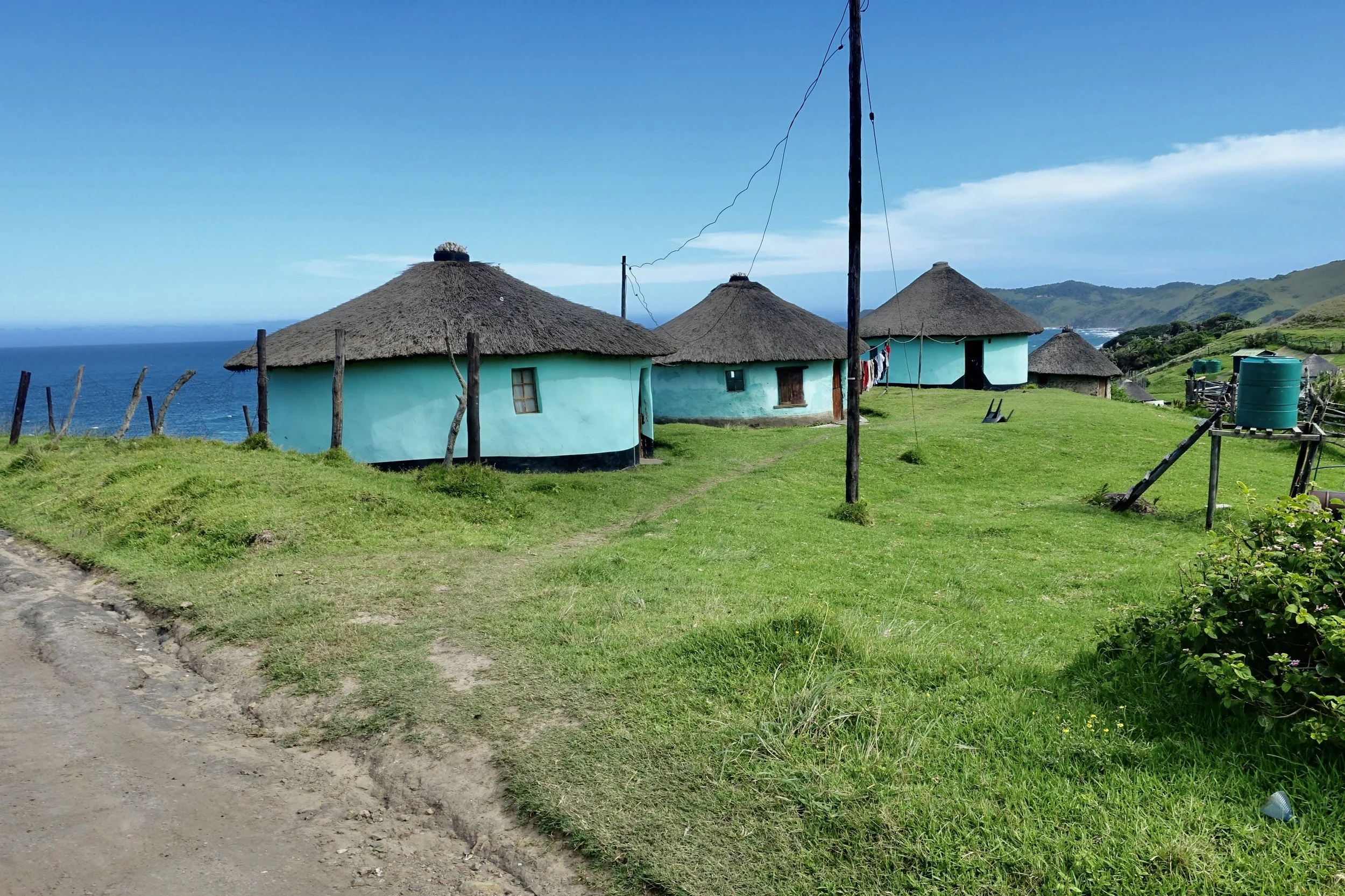 A scenic view of traditional round thatched-roof huts on a grassy hillside overlooking the ocean, with hills in the distance under a partly cloudy blue sky.