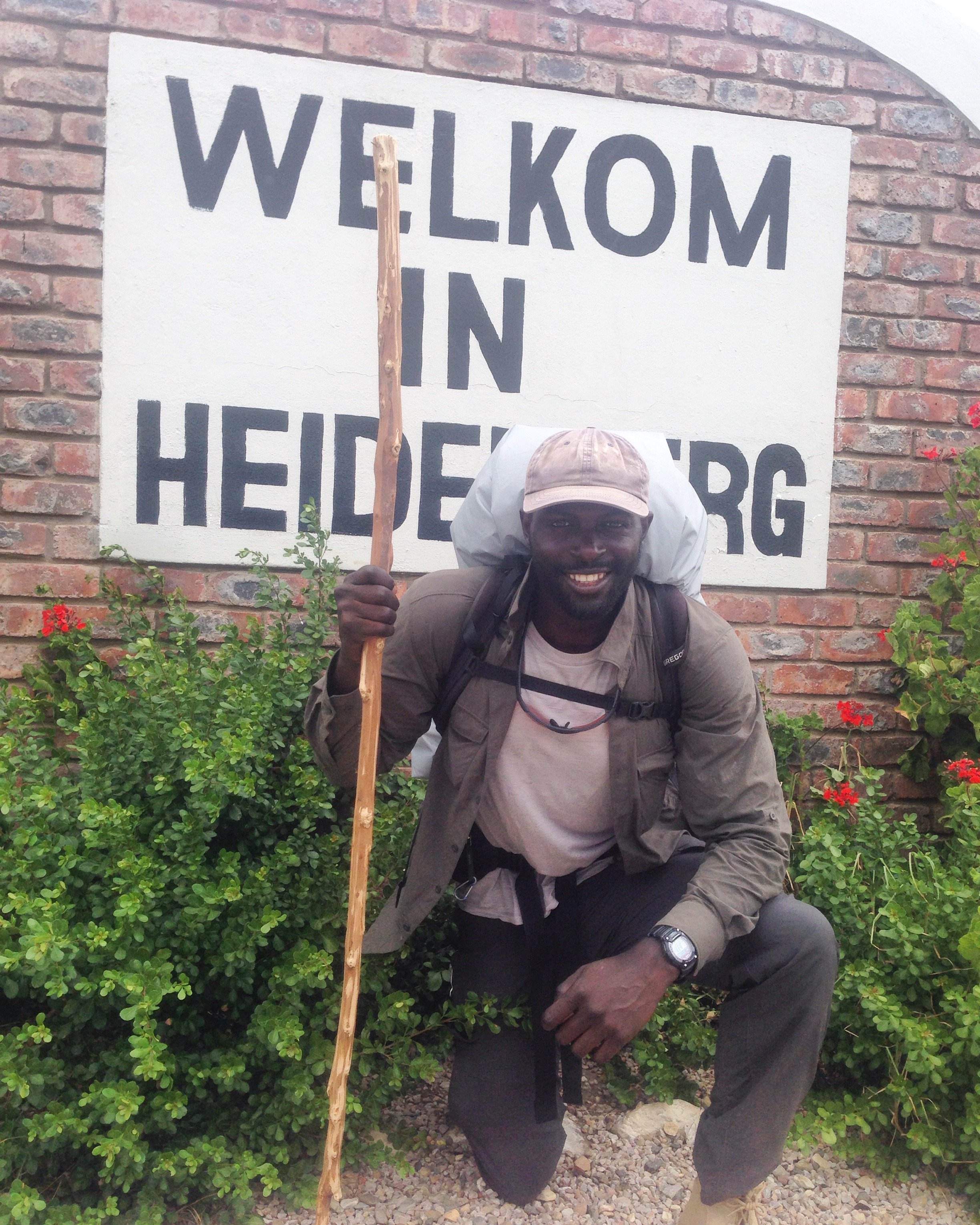 A man smiling, kneeling in front of a brick wall with a sign that reads 'Welkom in Heidelberg,' holding a walking stick, wearing outdoor gear, a backward cap, and a backpack.